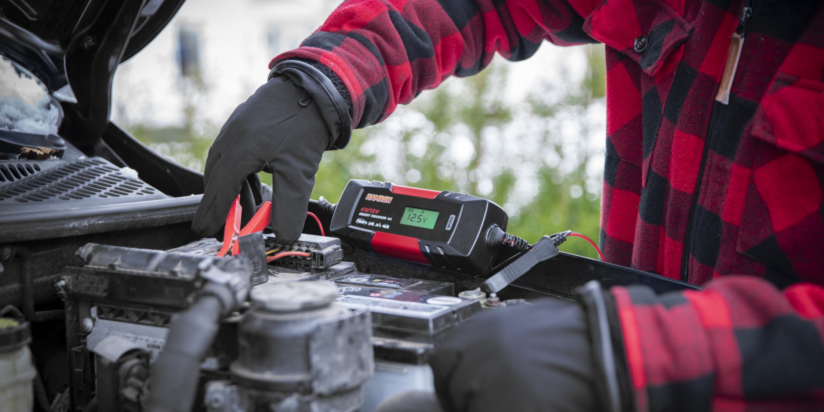 En person som bär handskar och en rödrutig jacka använder en HAMRON 6V/12V SMART CHARGER 4A för att ladda ett bilbatteri.