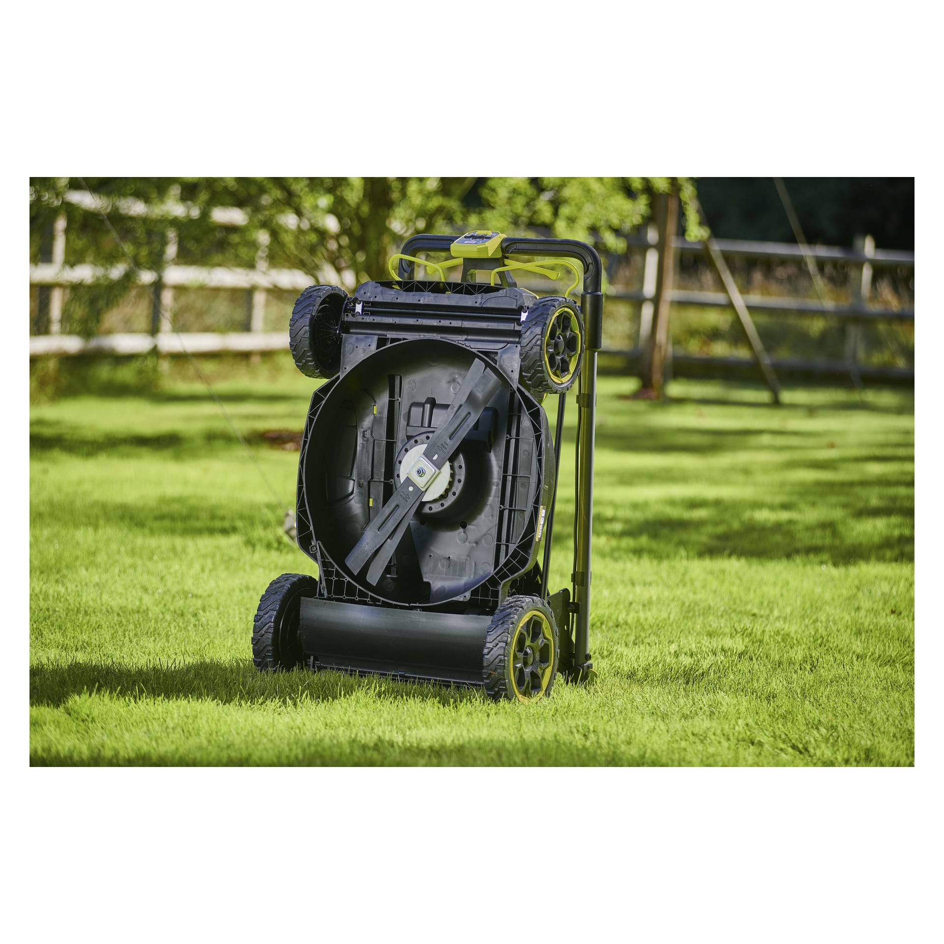 Ryobi Max Power gräsklippare RY36LMXSP53B-160 står upprätt på sidan i en gräsbevuxen trädgård, undersidan och bladet synliga, med staket och träd bakom.