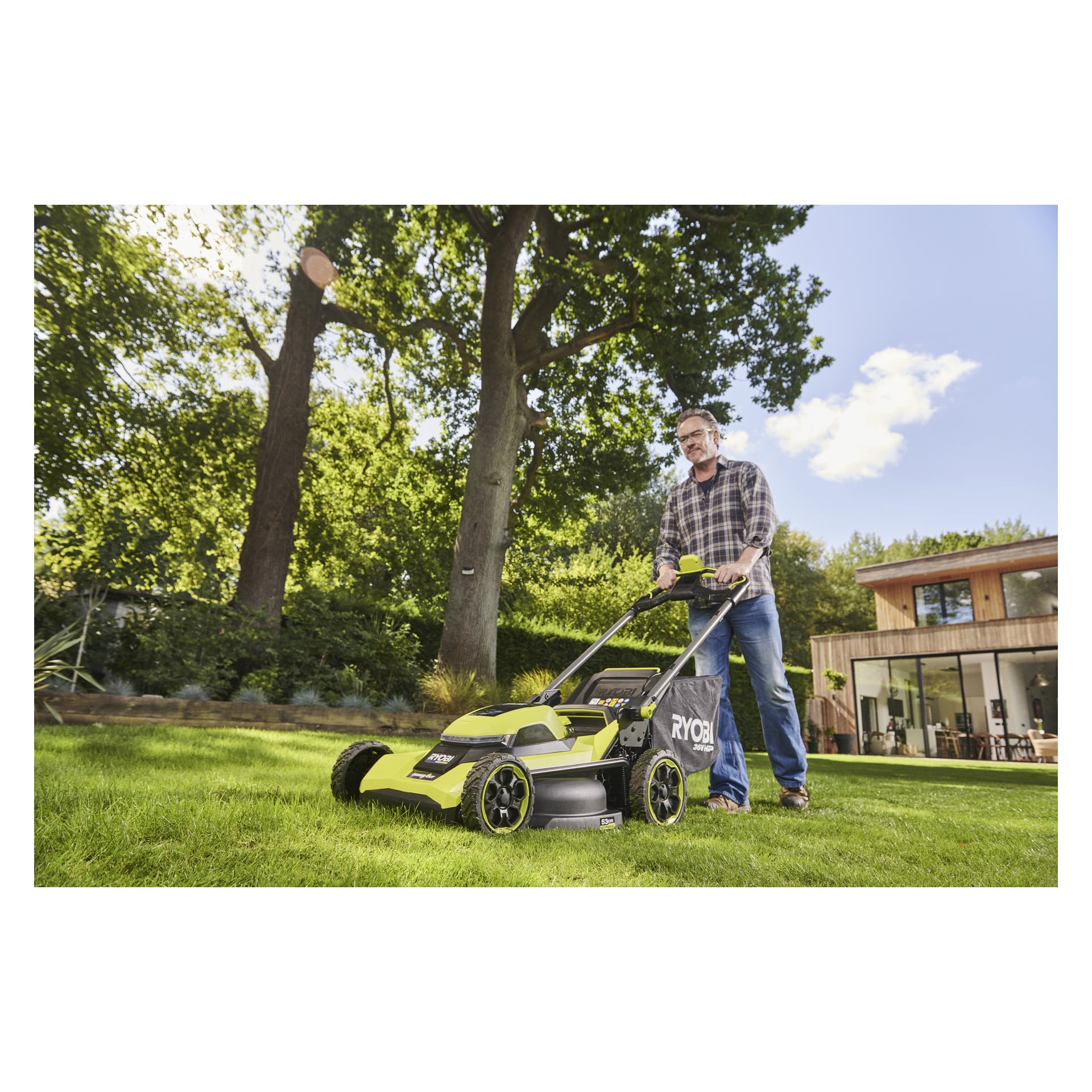 En man med glasögon och rutig skjorta använder en Ryobi Max Power-batteri gräsklippare på soligt gräs, med träd och ett modernt hus bakom.