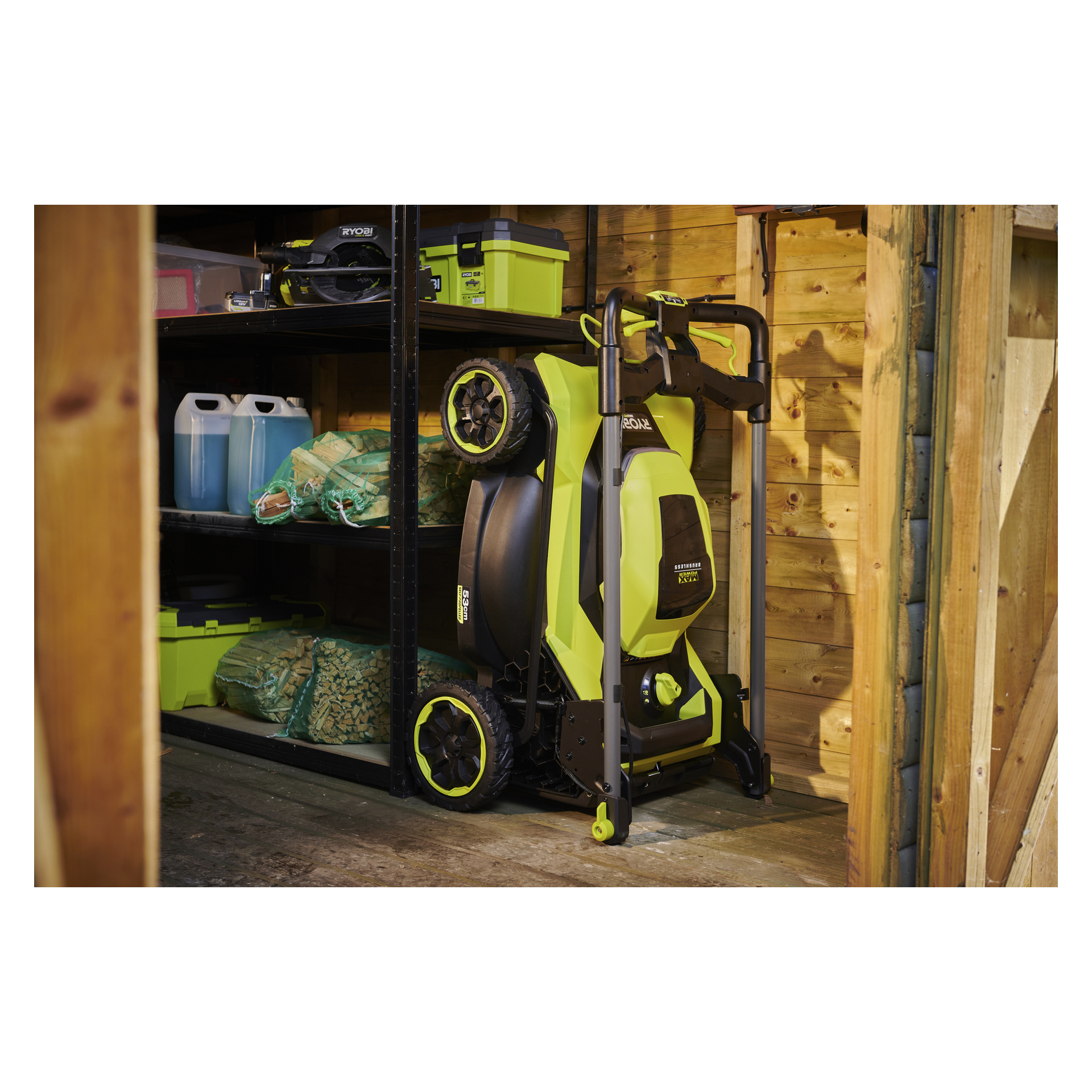 En grön och svart Ryobi Max Power-gräsklippare står upprätt bredvid metallhyllor i ett skjul med verktygslådor, blå kannor och trädgårdsväskor.