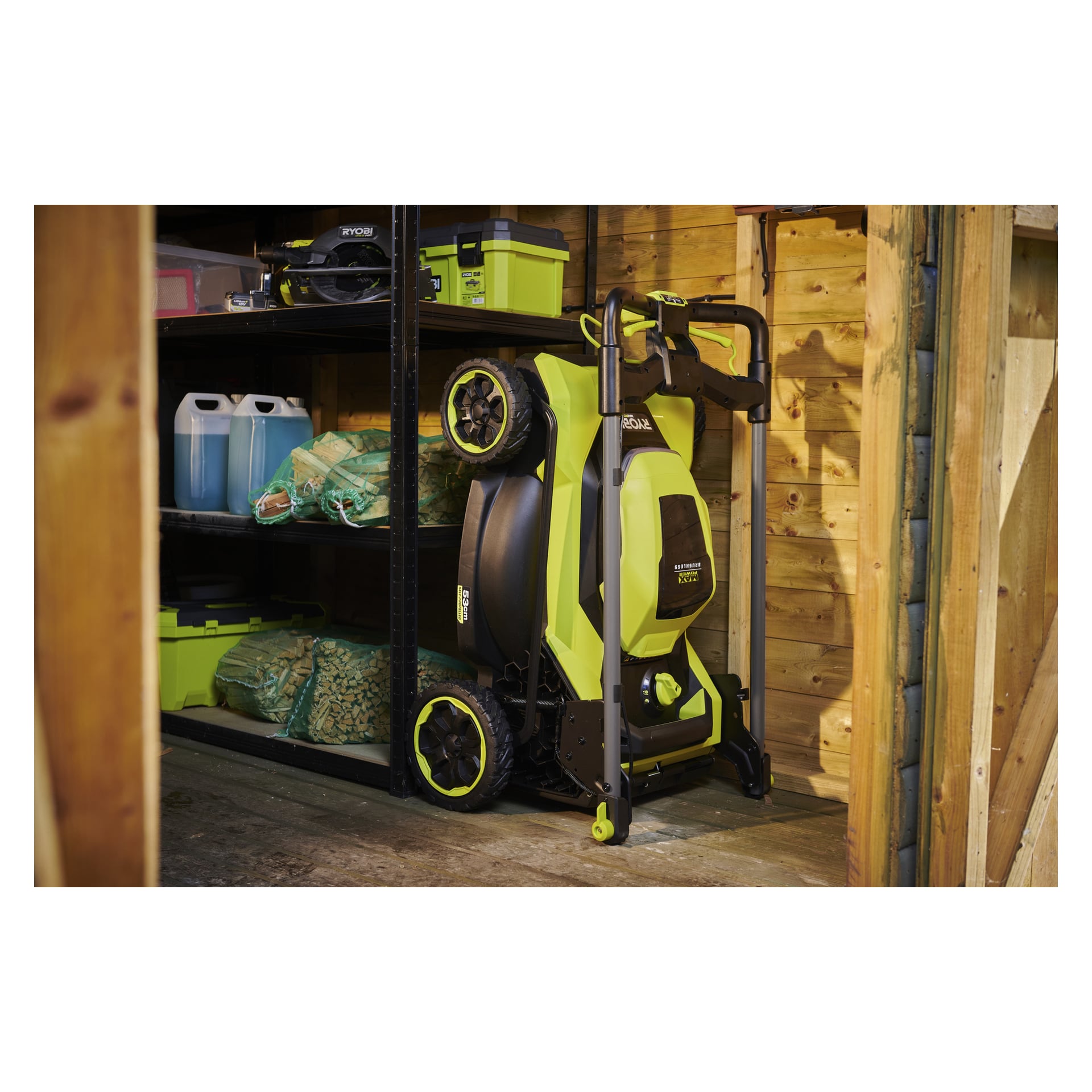 En grön och svart Ryobi Max Power-gräsklippare står upprätt bredvid metallhyllor i ett skjul med verktygslådor, blå kannor och trädgårdsväskor.