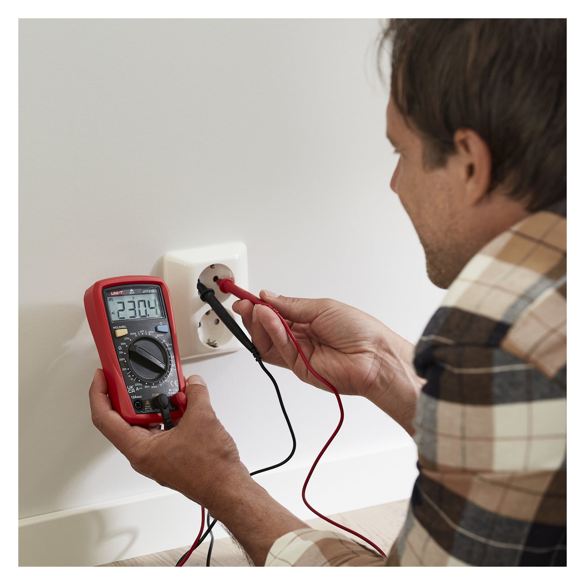 Någon i rutig skjorta håller sonder i ett vägguttag med hjälp av en digital multimeter som visar en spänningsavläsning på 230,4.