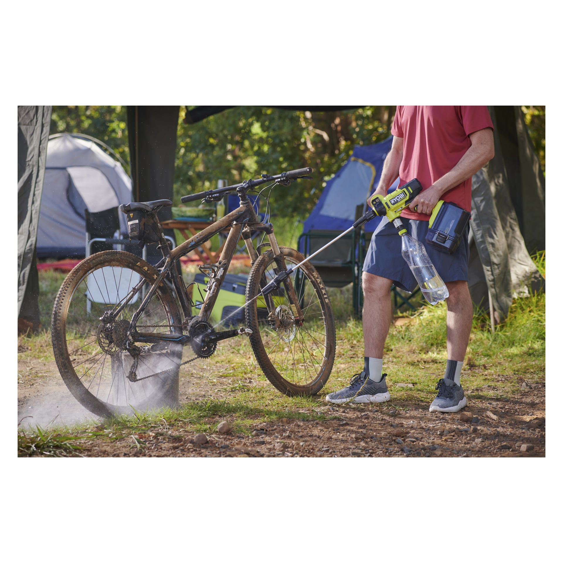 Iklädd en röd skjorta och blå shorts rengör en lerig cykel med en portabel högtryckstvätt nära tält och campingutrustning.