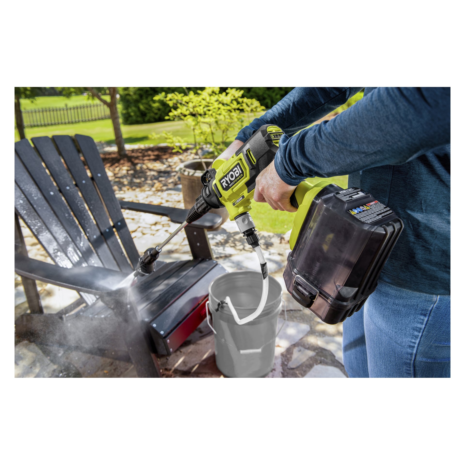 En person rengör en svart trästol utomhus med en Ryobi sladdlös högtryckstvätt, vattensprutande, med en grå hink nära utanför.
