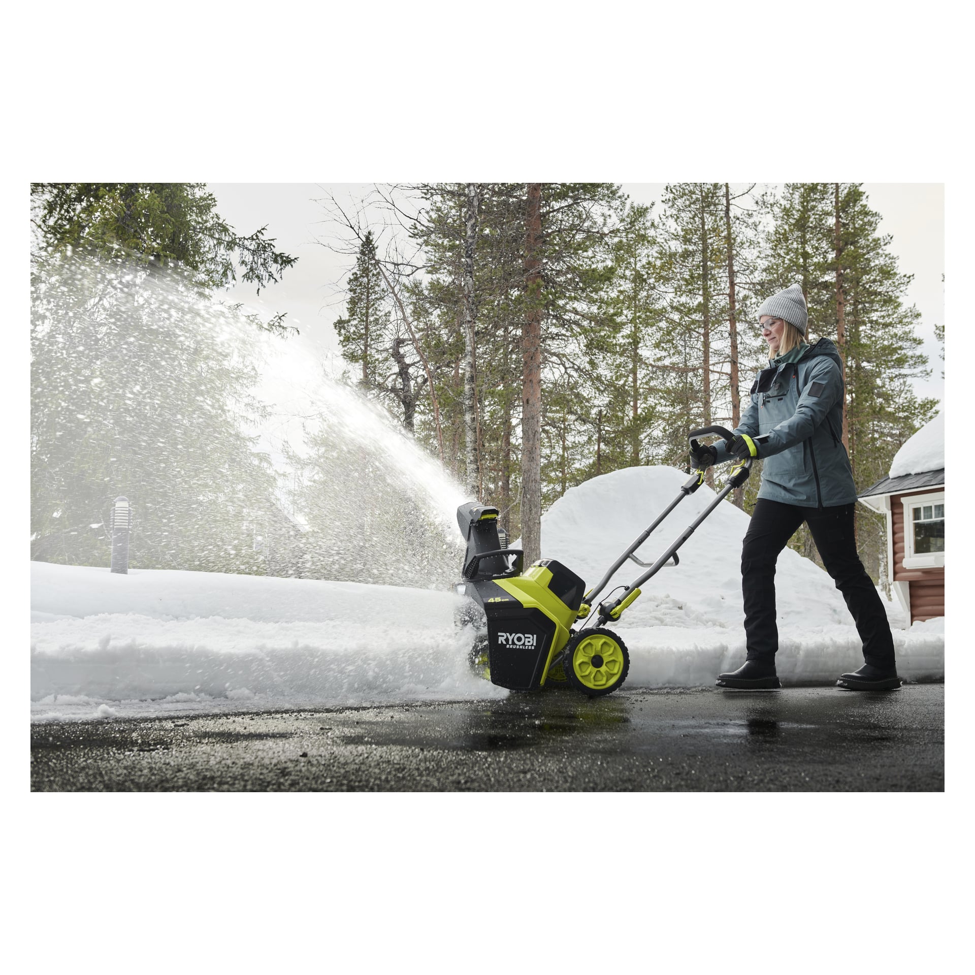 En person i vinterkläder kör en Ryobi-snöslunga på en uppfart och skickar undan snön, med träd och ett hus bakom sig.