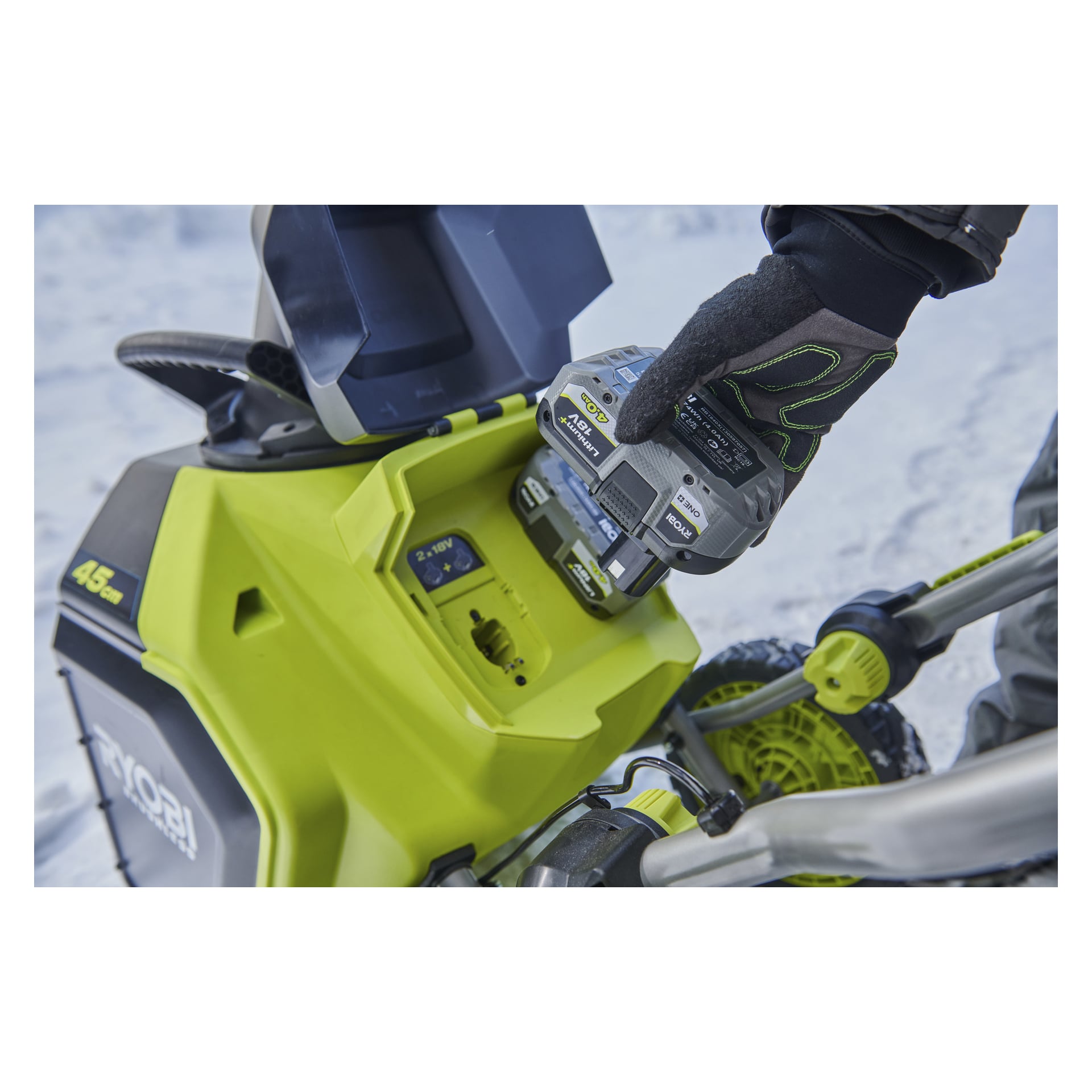 En handske skjuter in ett Ryobi 40V litiumbatteri i en grön och svart elektrisk snöslunga, snötäckt mark i bakgrunden.