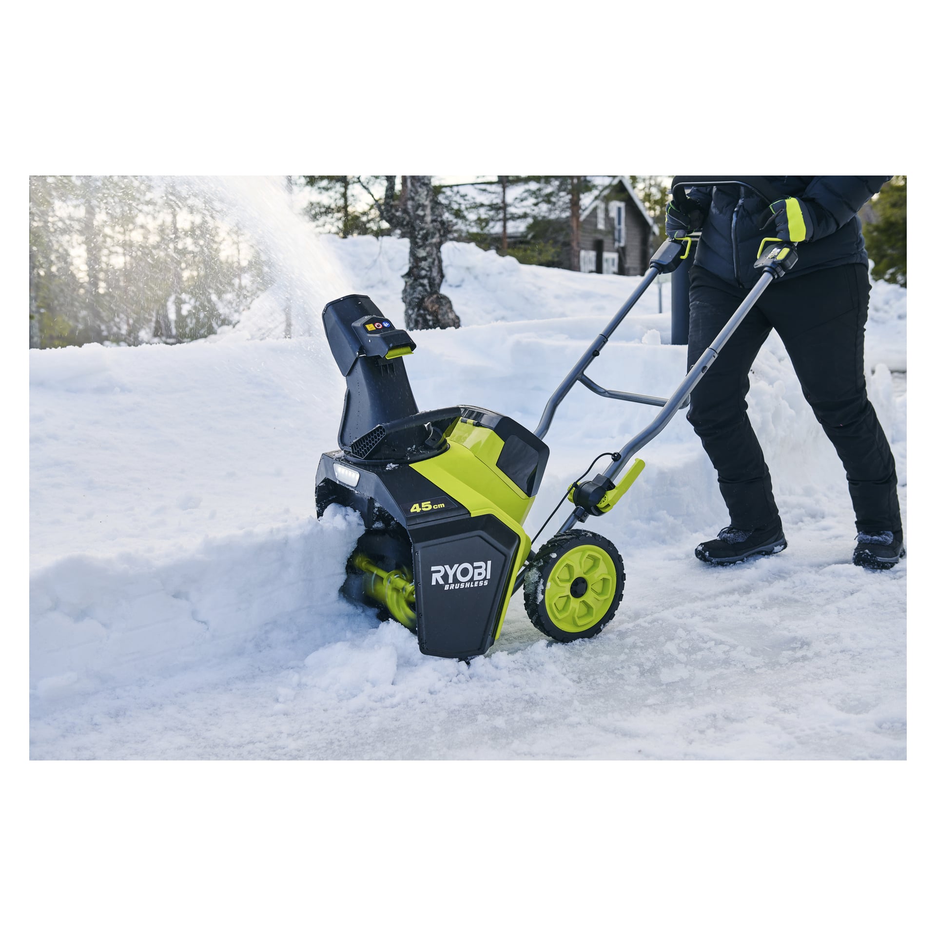Insvept i vinterkläder kör en person en Ryobi elektrisk snöslunga genom djup snö, träd och ett hus bakom sig.