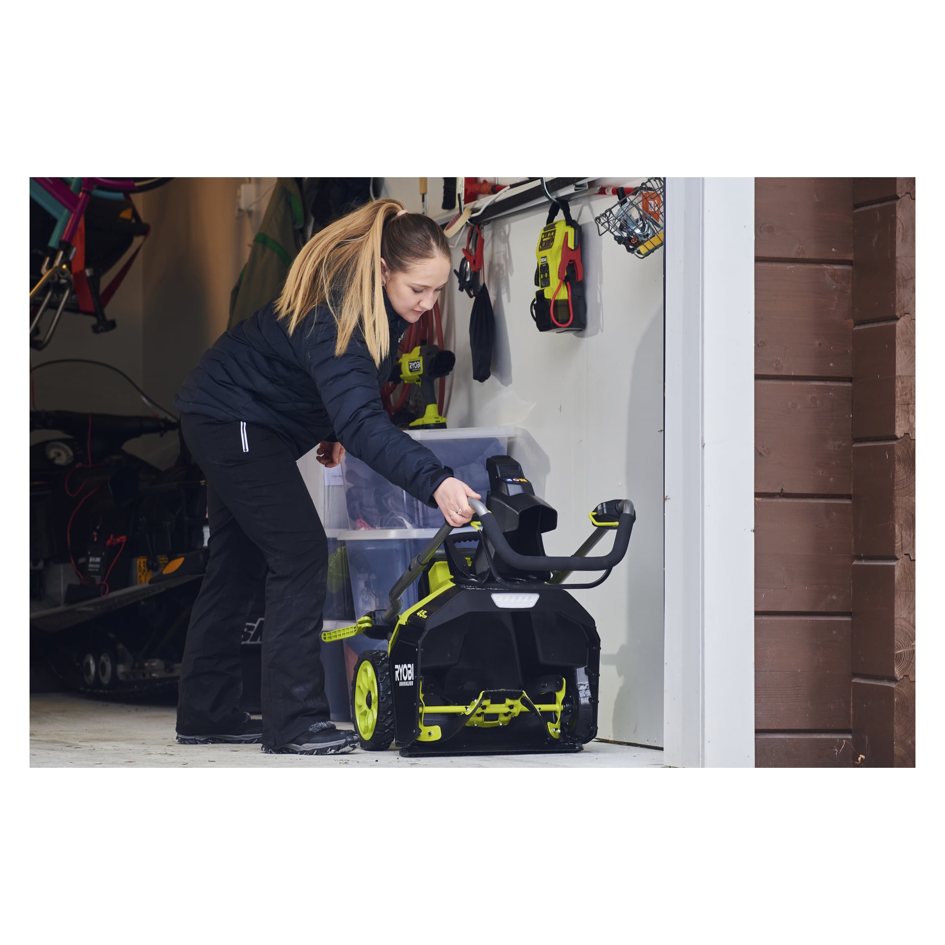 En kvinna i svart jacka och byxor förvarar en grön och svart Ryobi-snöslunga i ett garage med verktyg och utrustning på hyllorna.