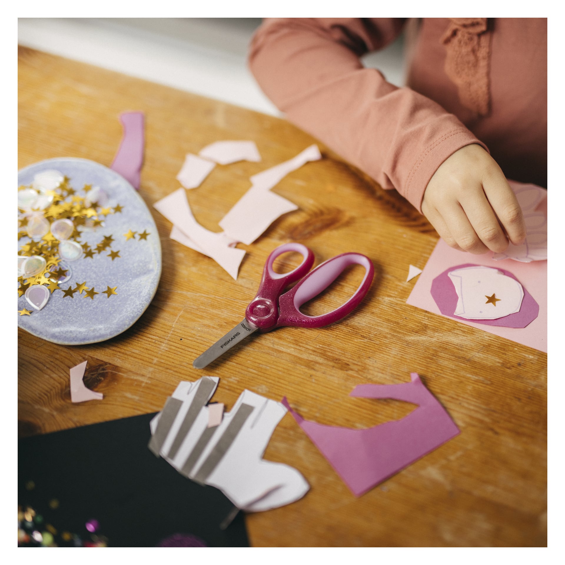 Et barns hånd strekker seg etter rosa papirformer blant hobbyartikler som saks, gullstjernepaljetter og utskåret papir på et trebord.