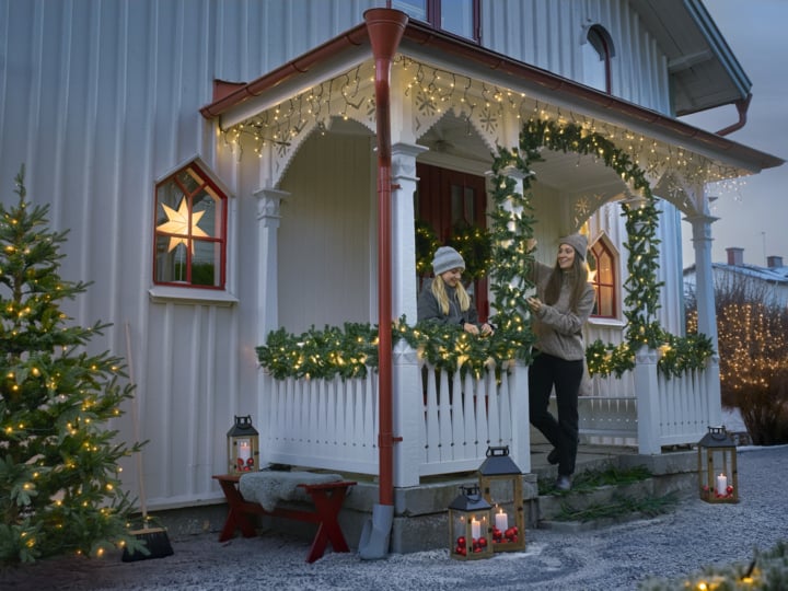 Eine festliche Veranda mit Lichtern und Girlande. Zwei Menschen unterhalten sich lächelnd zwischen Laternen, einem Weihnachtsbaum und schneebedecktem Schmuck.