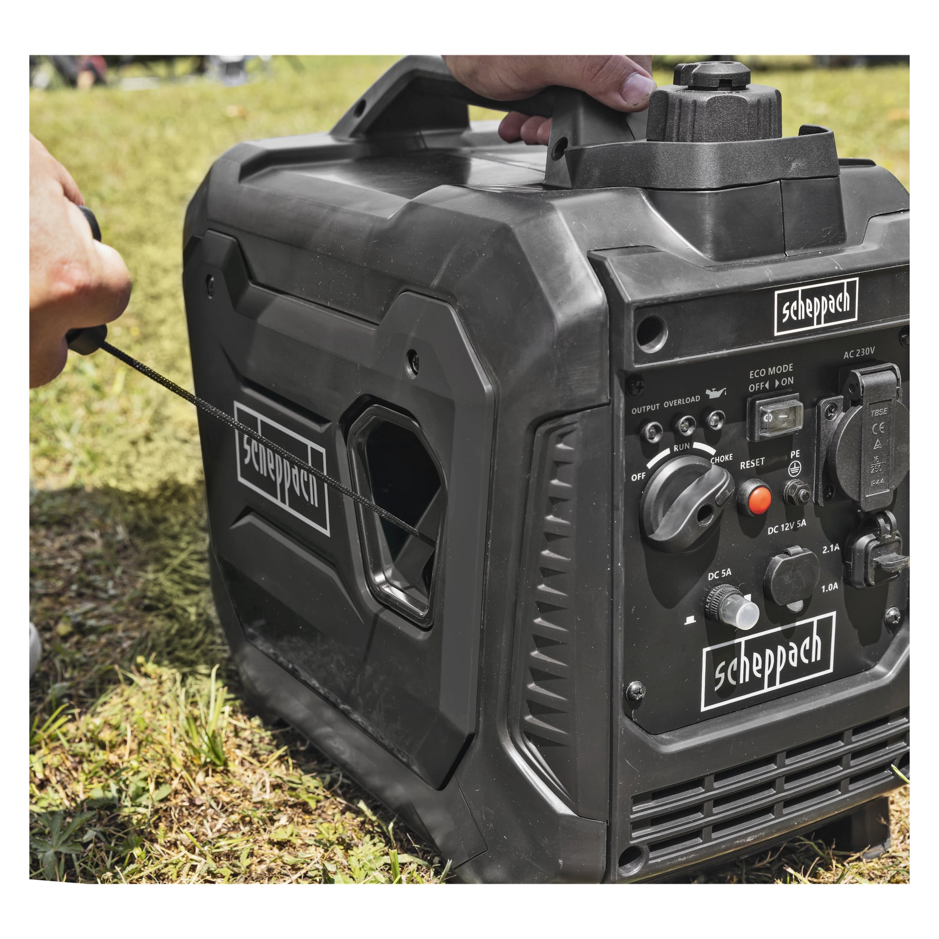 Någon startar en svart bärbar Scheppach-generator på gräs, med en panel med knappar, knoppar och uttag.