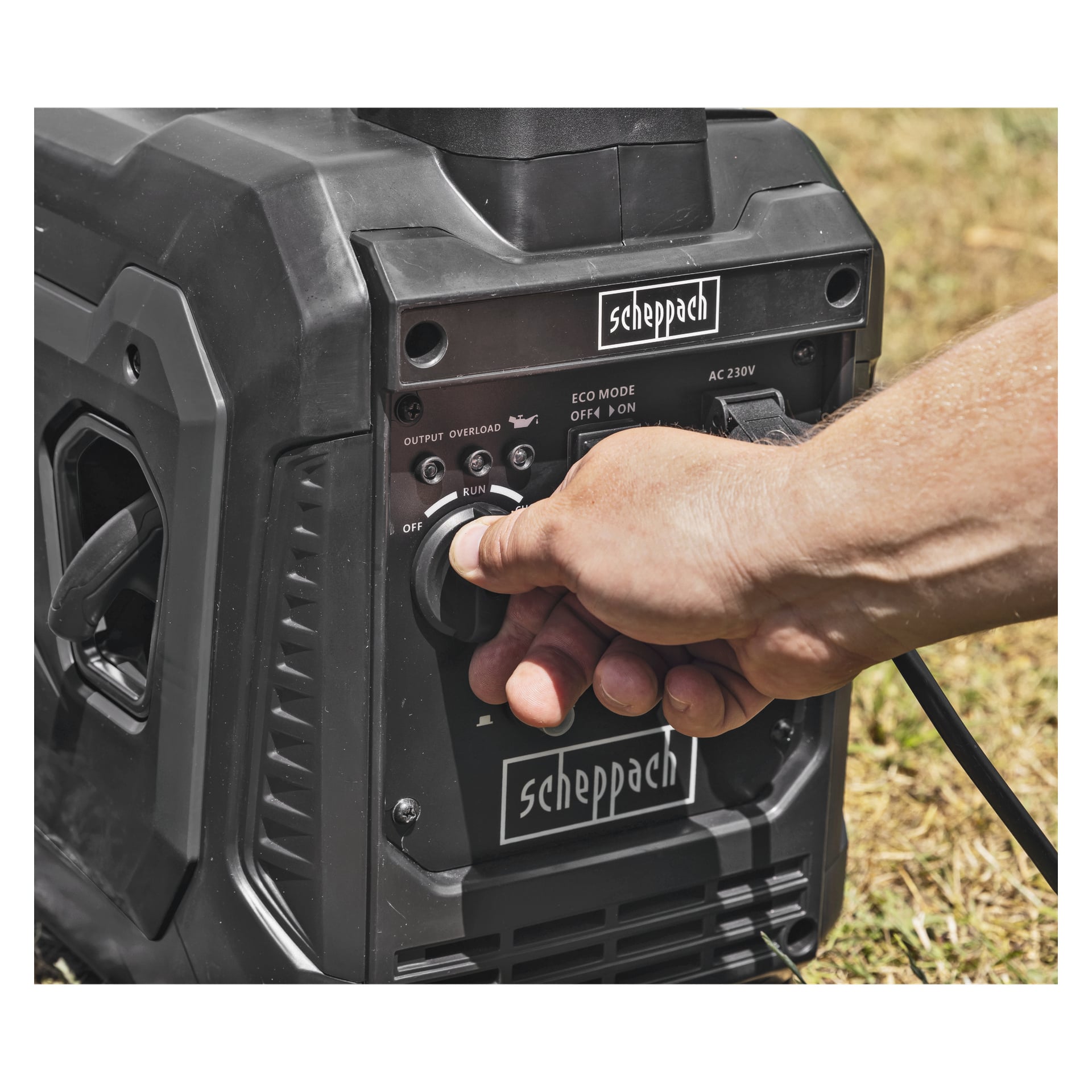 En hand justerar kontrollvredet på en svart Scheppach-generator utomhus, omgiven av gräs.