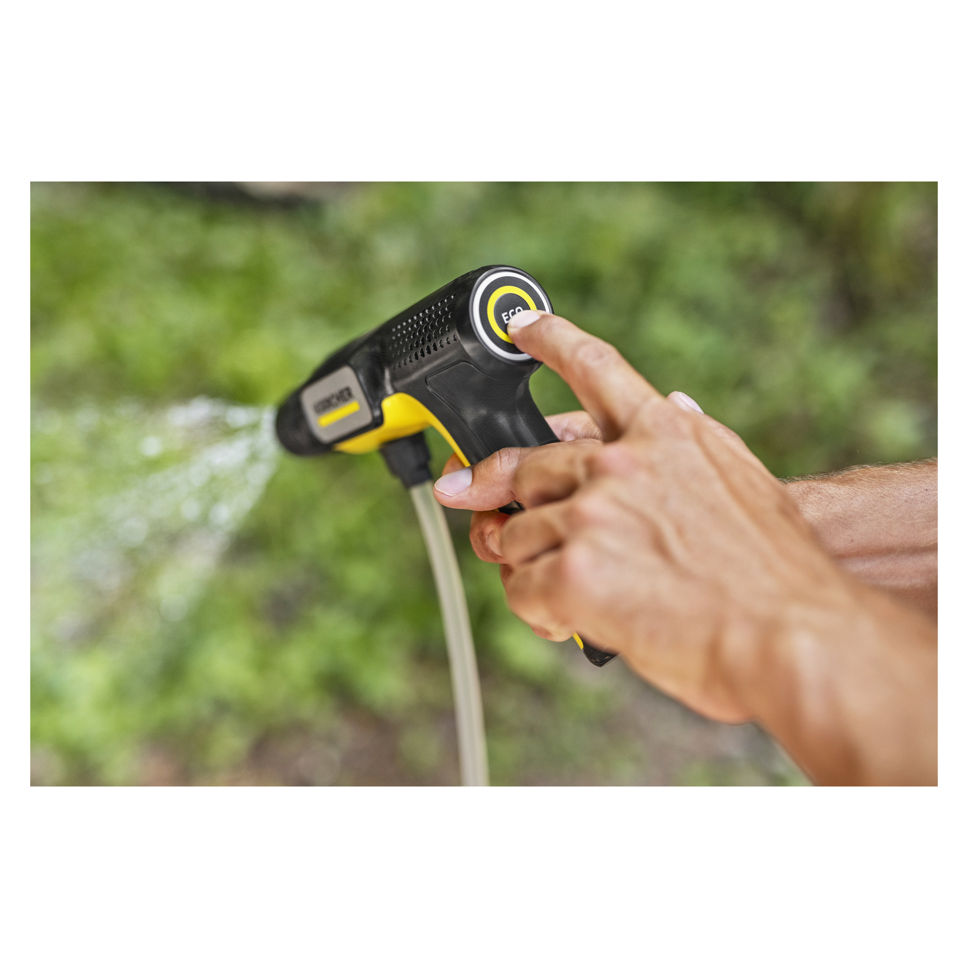 En hand trycker på sprayknappen på ett svart och gult munstycke för trädgårdsslangen och sprutar vatten mot en suddig grön bakgrund.