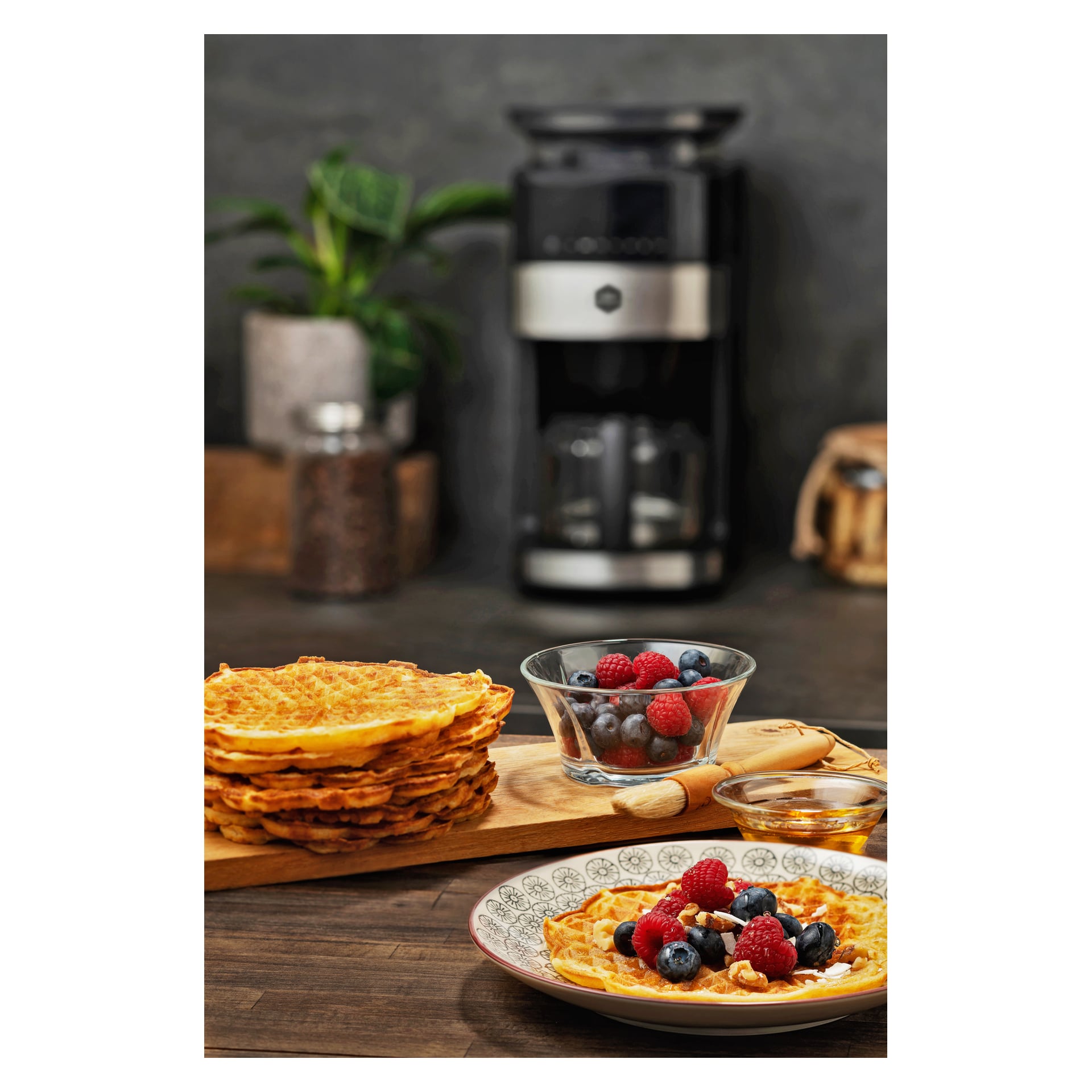 Vafler stablet på et skjærebrett, en bolle med blandede bær, vafler med bær på en tallerken og en kaffetrakter på benken.