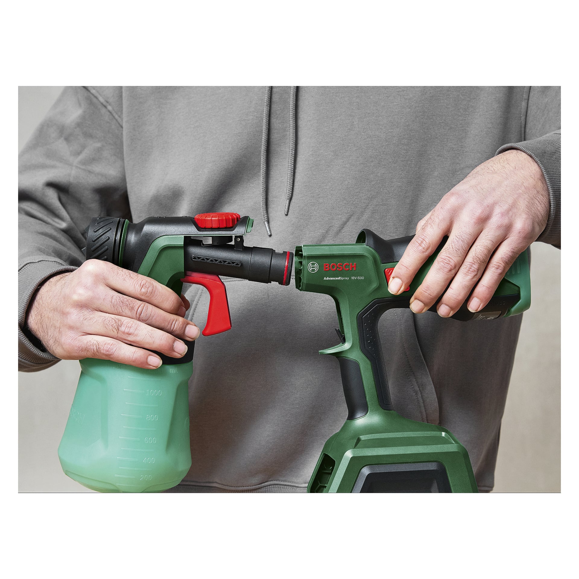 Iklädd en grå hoodie ansluter en person en grön och svart Bosch spraytillsats till sitt verktyg; Den halvtransparenta flaskan visar mått.