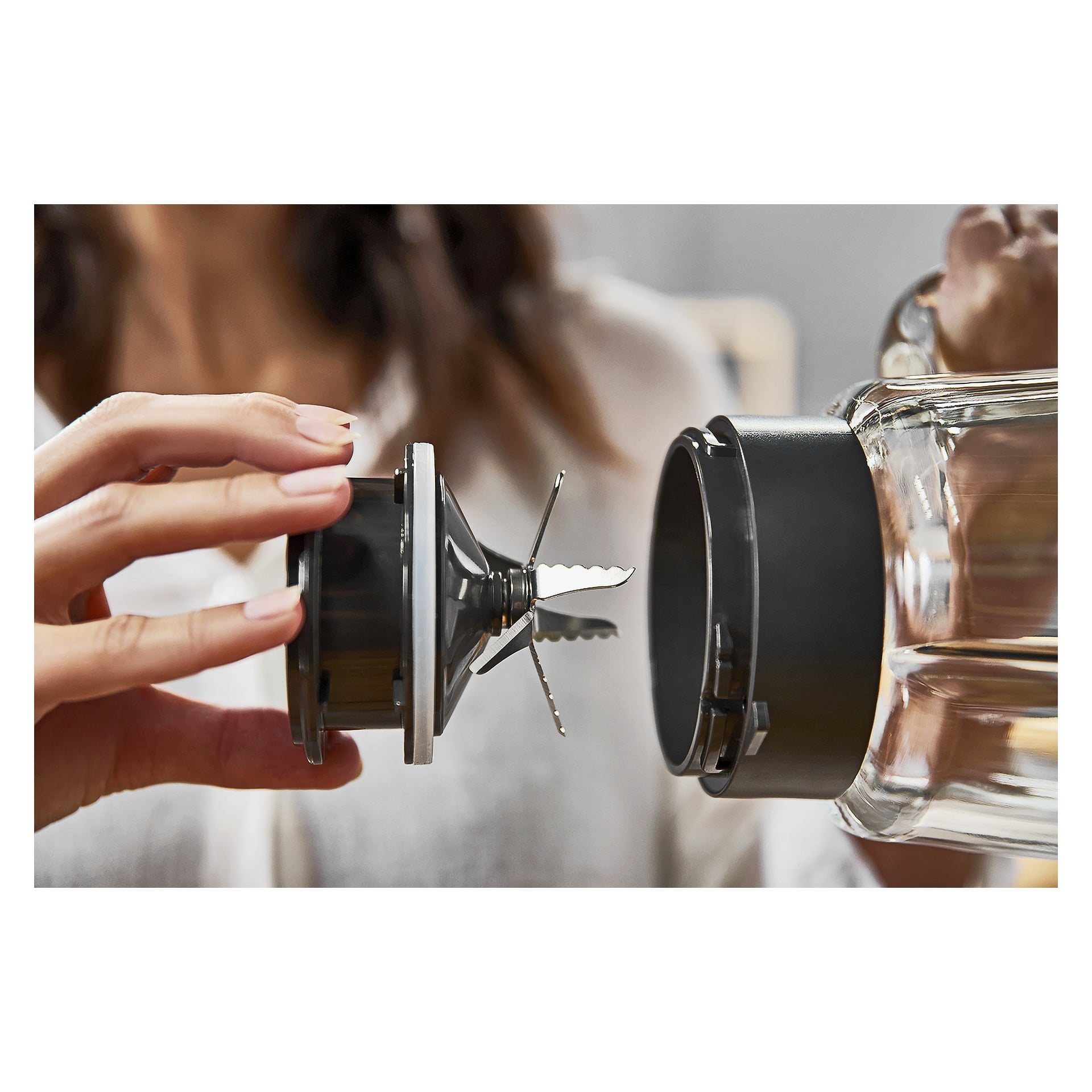 En hand håller ett svart mixerblad och placerar det så att det passar in i den öppna basen på en mixerburk i glas, med en suddig bakgrund.