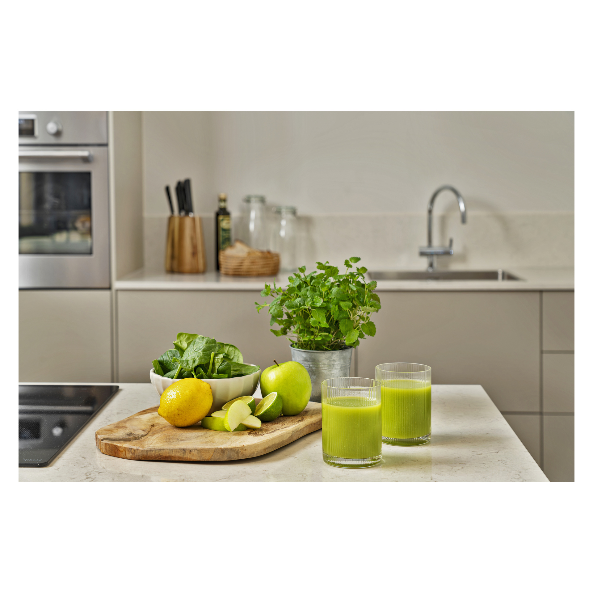 Två glas grön juice, citron, äpple, spenat på en bräda, skål med bladgrönsaker och krukväxt på en köksbänk.