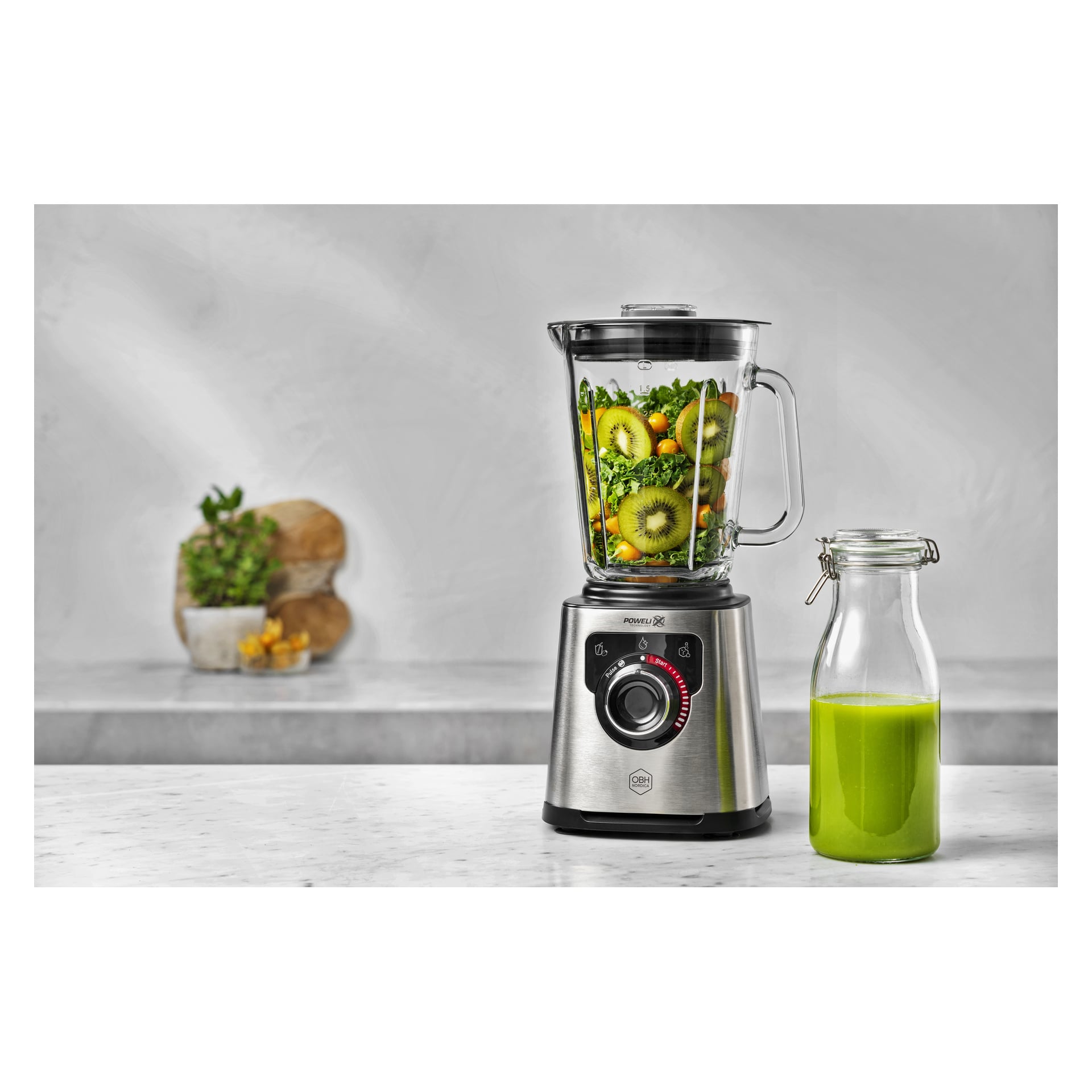 På en bänk står en mixer fylld med frukt och grönsaker bredvid en förseglad burk med grön juice; bröd och en växt i ryggen.