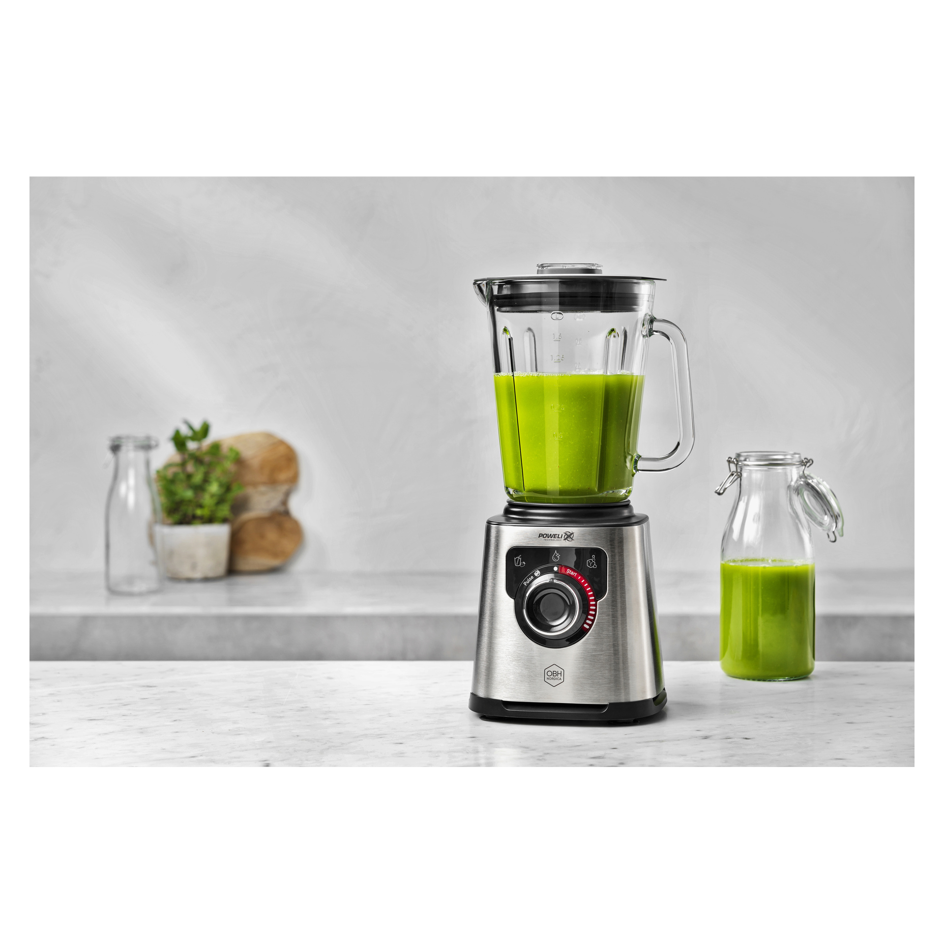 En mixer i rostfritt stål med grön vätska står på en bänk bredvid en matchande glasflaska; Bröd och en liten växt ligger bakom.