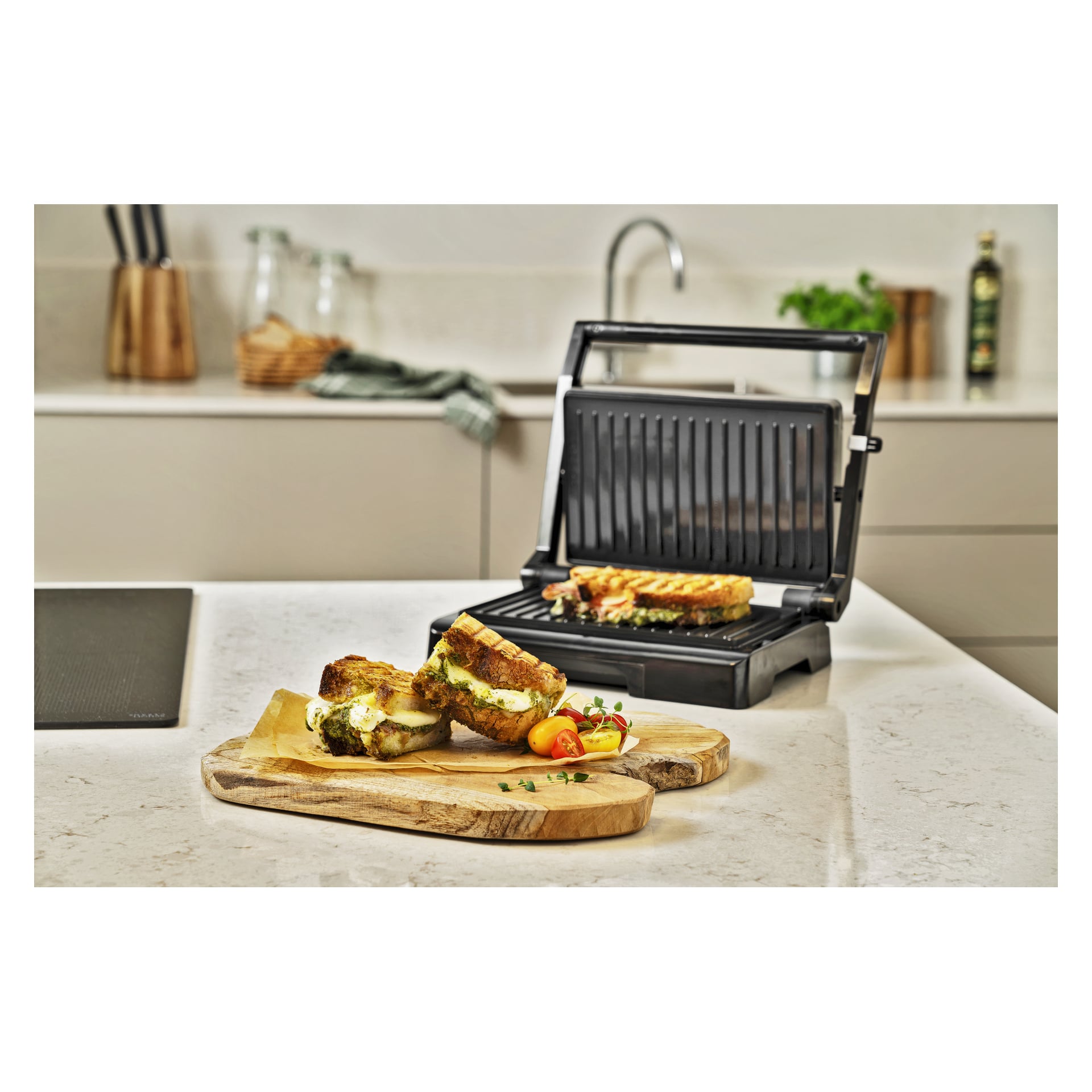 En paninipress grillar en smörgås på bänken; I närheten står två smälta pestosmörgåsar på en träbräda med tomater och örter.