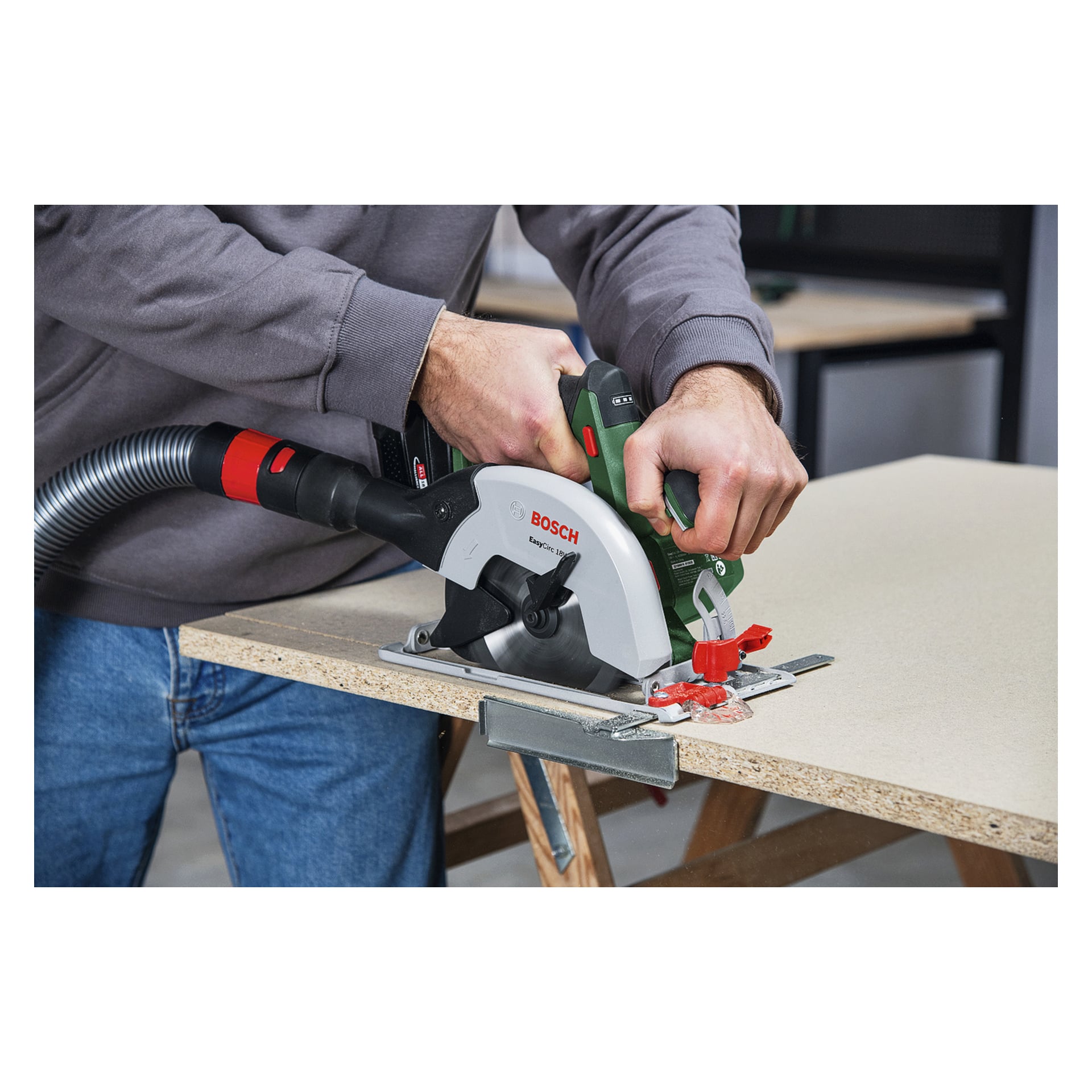 En person griper en Bosch sirkelsag med begge hender og skjærer sponplater på en arbeidsbenk mens en vakuumslange samler støv.