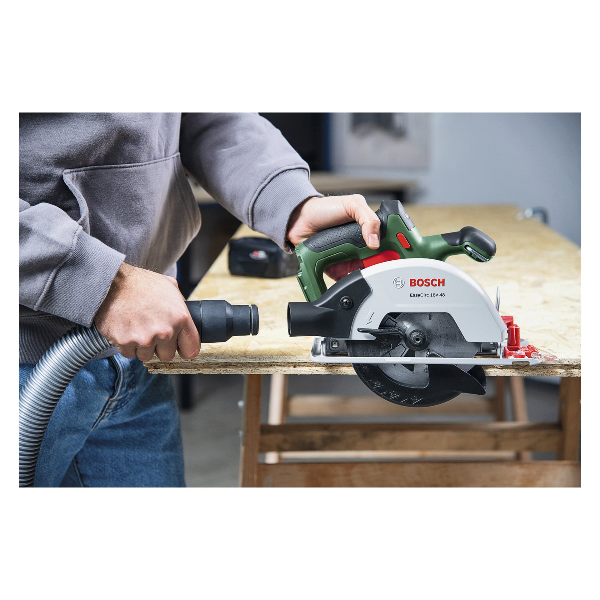 Iført en grå hettegenser kutter en person en treplate på en arbeidsbenk med en Bosch sirkelsag, og holder vakuumslangen for støv.