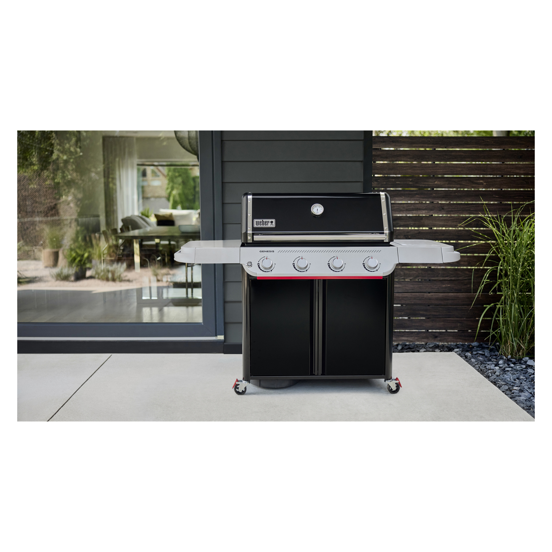 En svart och silverfärgad gasgrill med fyra knoppar och två sidohyllor står på en uteplats nära ett mörkpanelat hus med glasdörr.