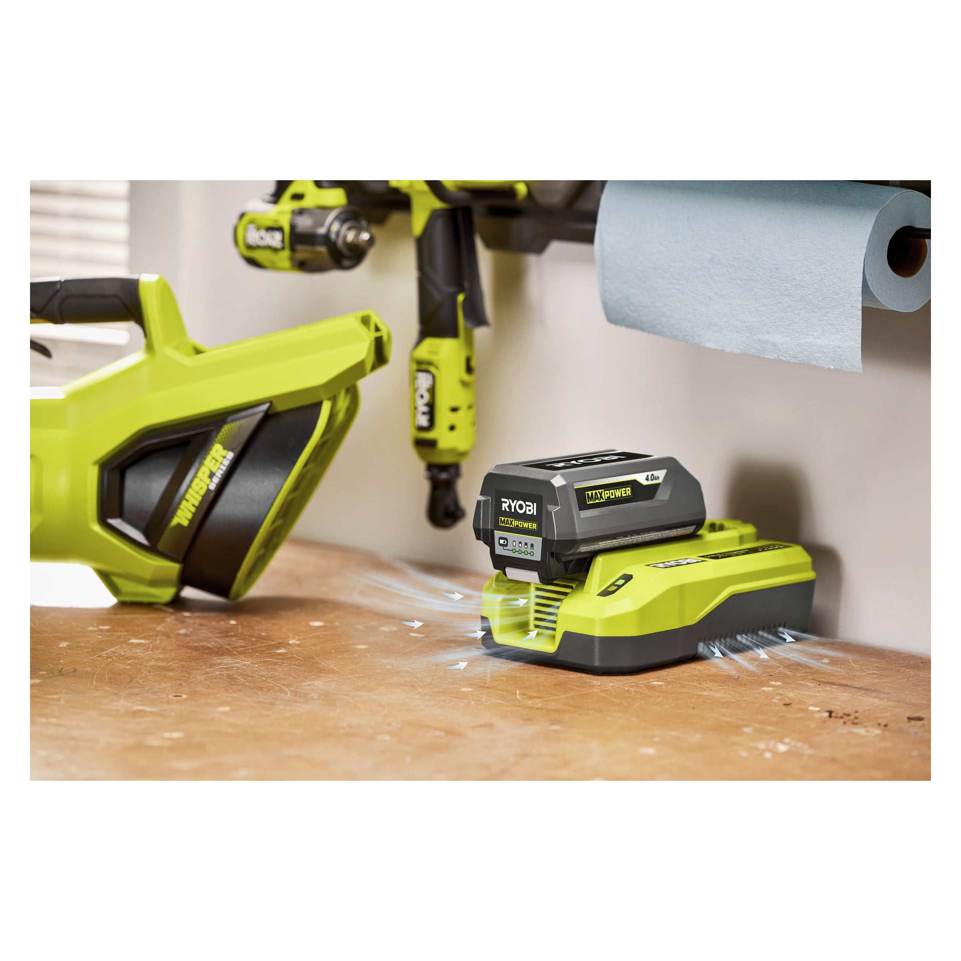 Ett Ryobi 40V litiumbatteri laddas i en grön laddare på en arbetsbänk av trä, med ett sladdlöst verktyg och en pappershandduksrulle monterade ovanför.