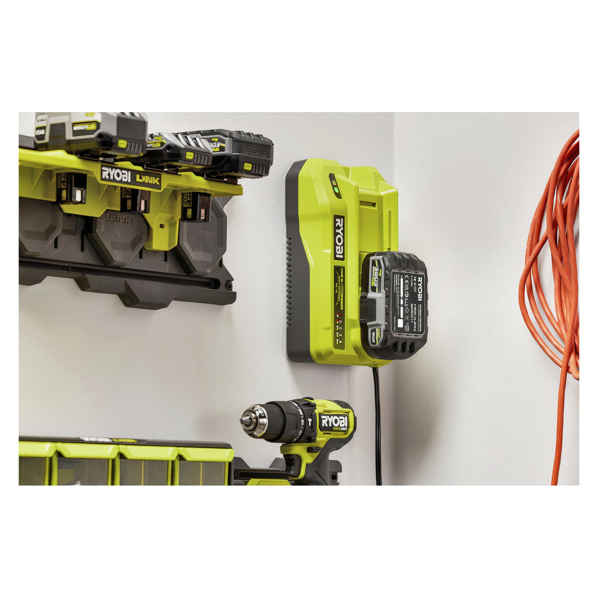 En Ryobi-laddare med batteri är väggmonterad, med en sladdlös borrmaskin och extra batterier i närheten på ett rack; Orange snöre hänger till höger.