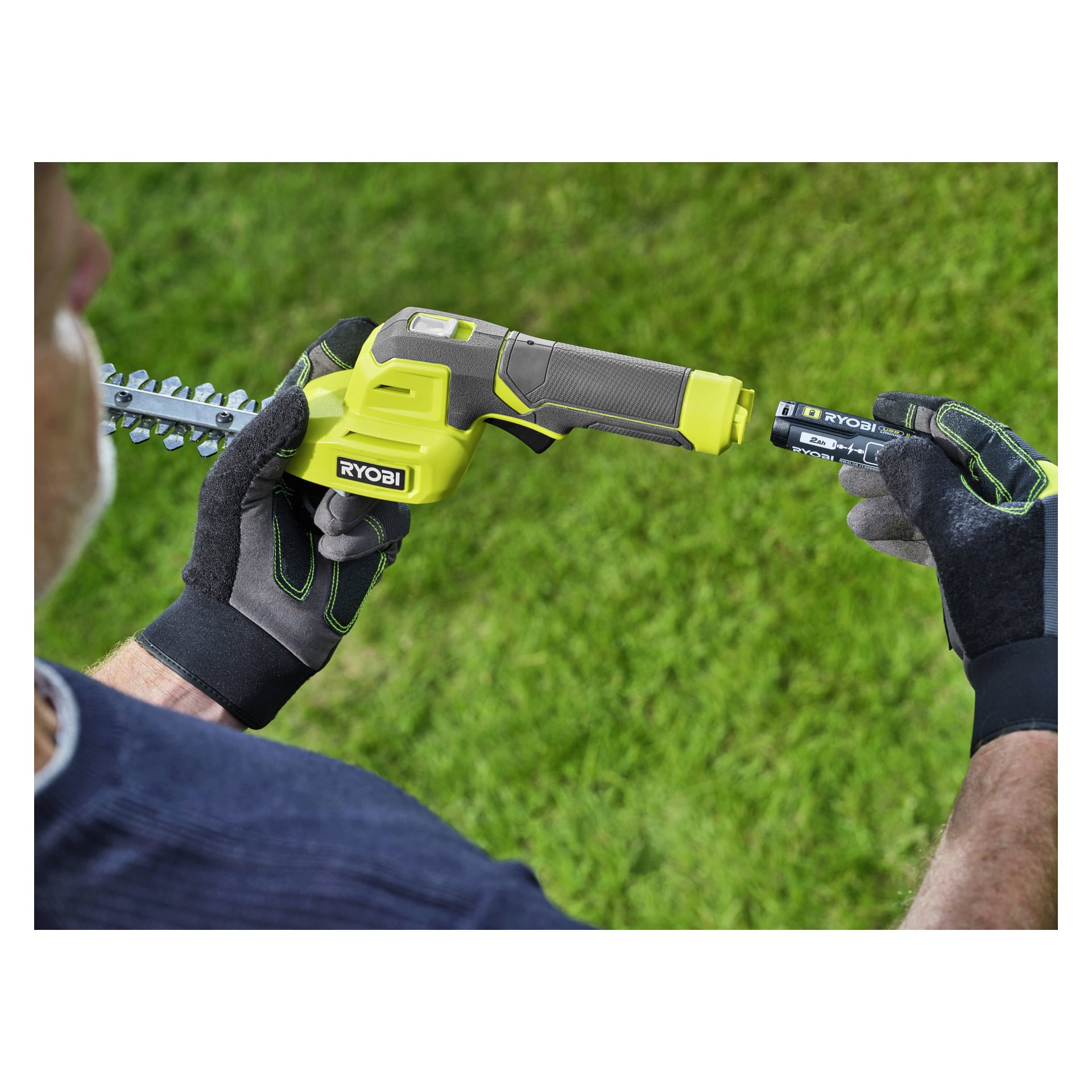 Handskbeklädda händer sätter in ett batteri i ett handhållet elverktyg från Ryobi, med grönt gräs i bakgrunden.