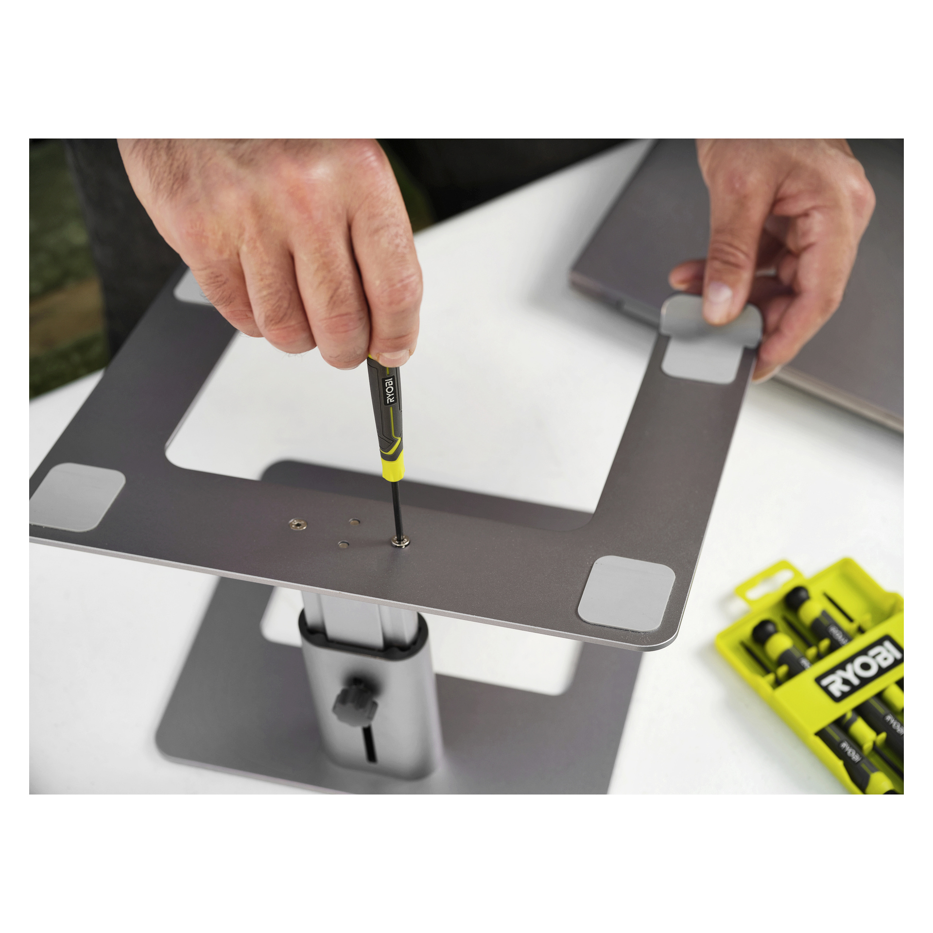 Någon fäster skruvar i ett metallstativ med en gul och svart Ryobi-skruvmejsel; ett Ryobi-batteripaket sitter i närheten.