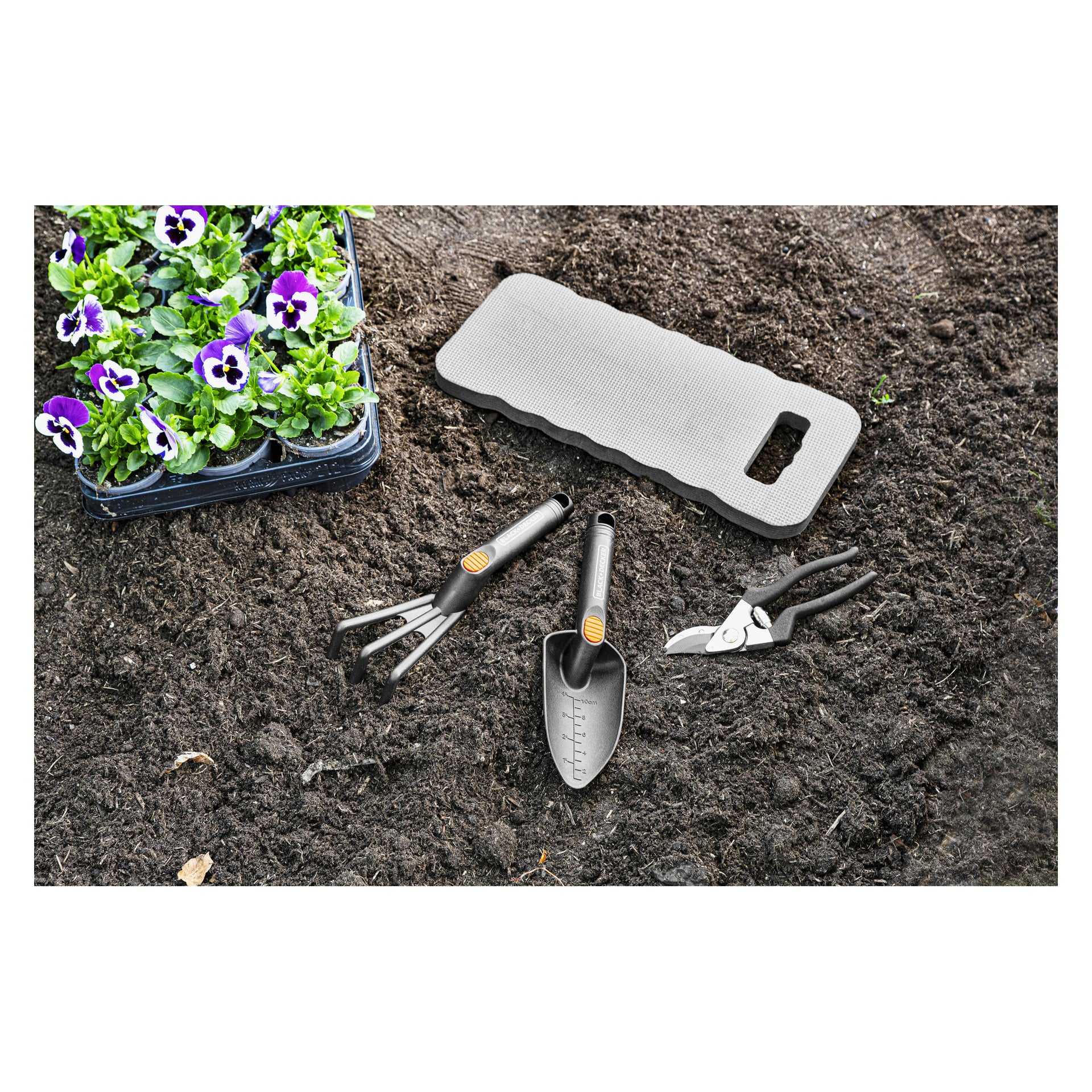 Lila och vita blommor i en bricka, grå knäböj, handkratta, murslev och sax placerade på mörk jord för trädgårdsarbete.