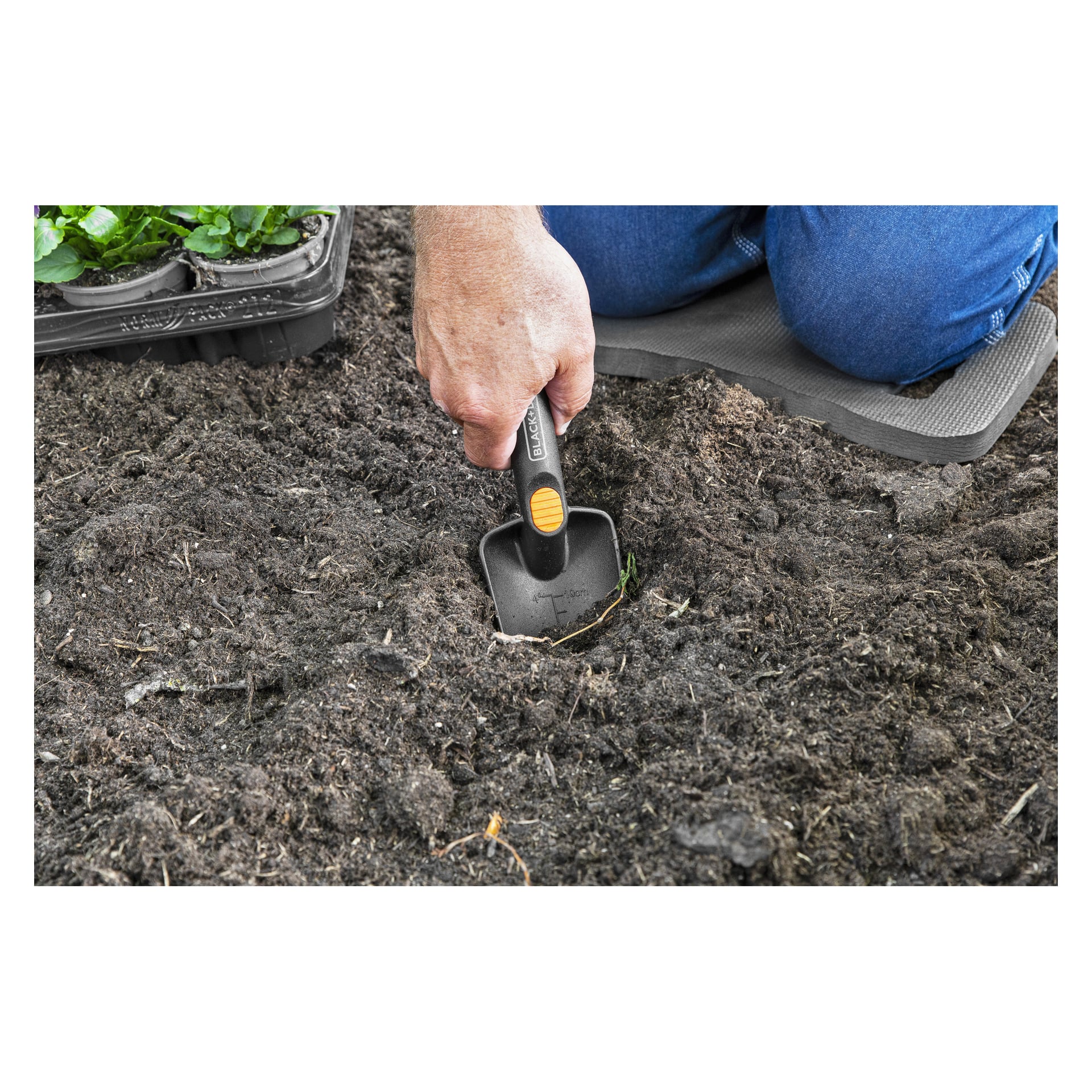 Knäböjande på en skumplatta gräver en person i mörk jord med en svart handspade; Plantor i en bricka står uppe till vänster.