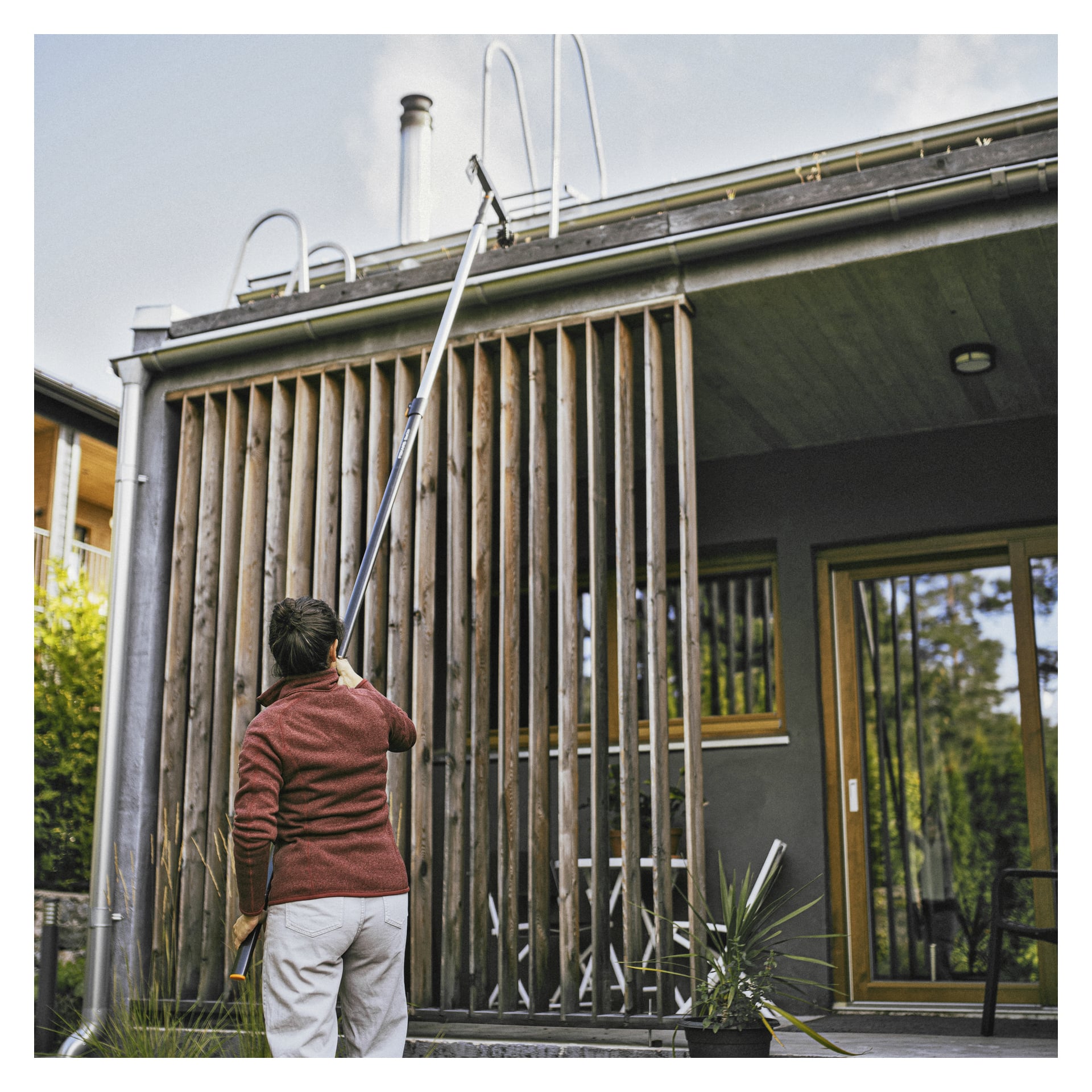 Mit einem roten Pullover benutzt man eine lange Stange, um an die Rinne auf einem modernen Haus mit Holzlatten und großen Glastüren zu gelangen.