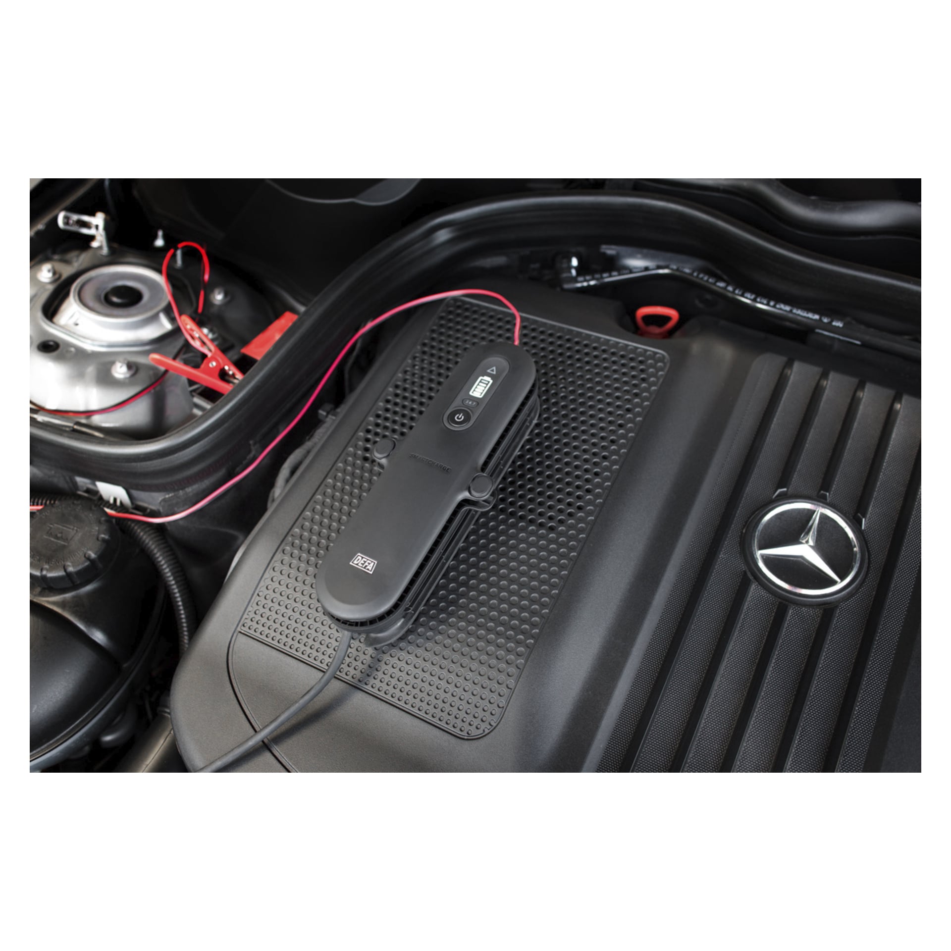 Närbild på en svart startmotorcykel kopplad till ett Mercedes-Benz bilbatteri, med röda och svarta kablar fastklämda på terminalerna.