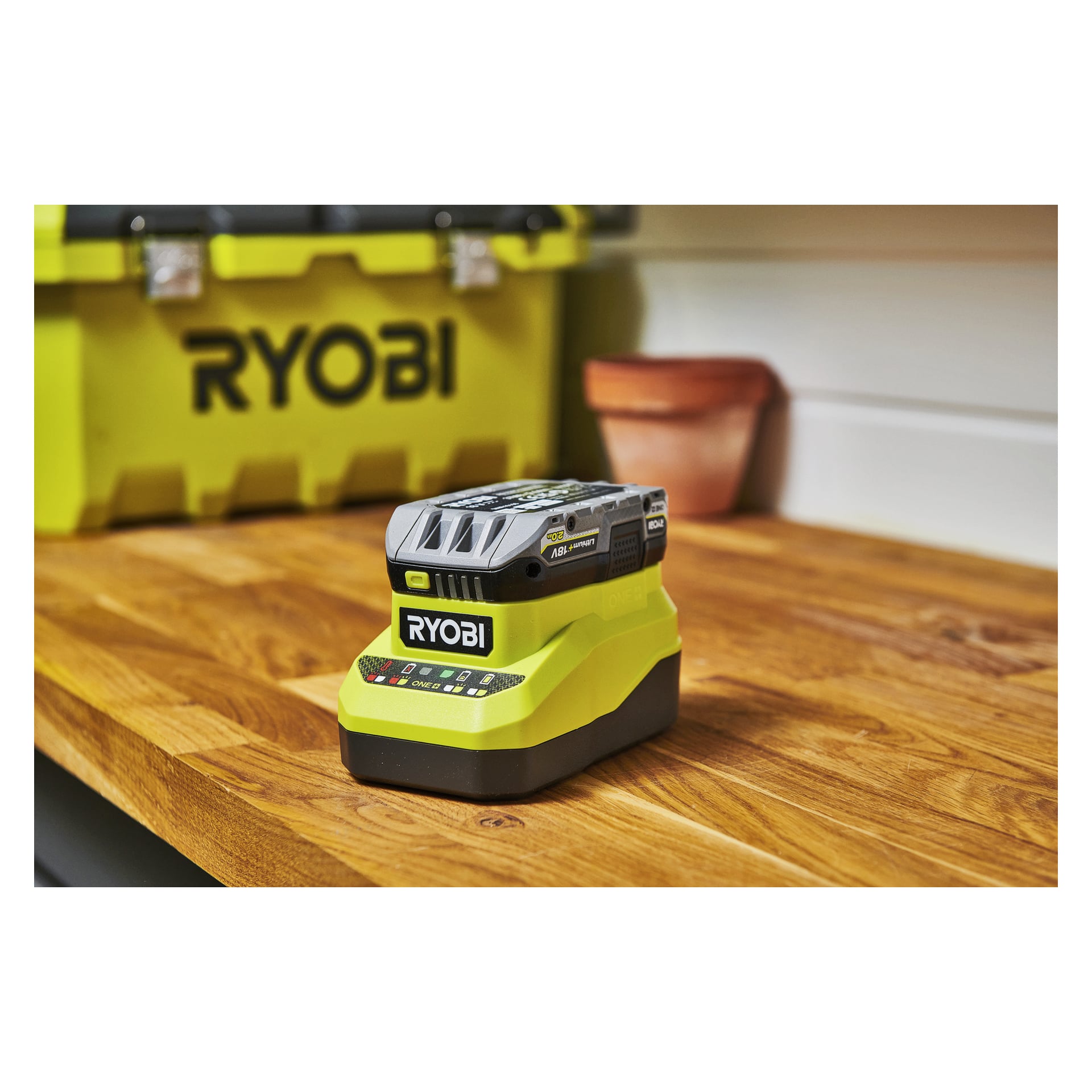 Ein Ryobi-Batterieladegerät steht auf einer hölzernen Werkbank, dahinter befindet sich eine Ryobi-Aufbewahrungsbox und ein kleiner Terrakottatopf.