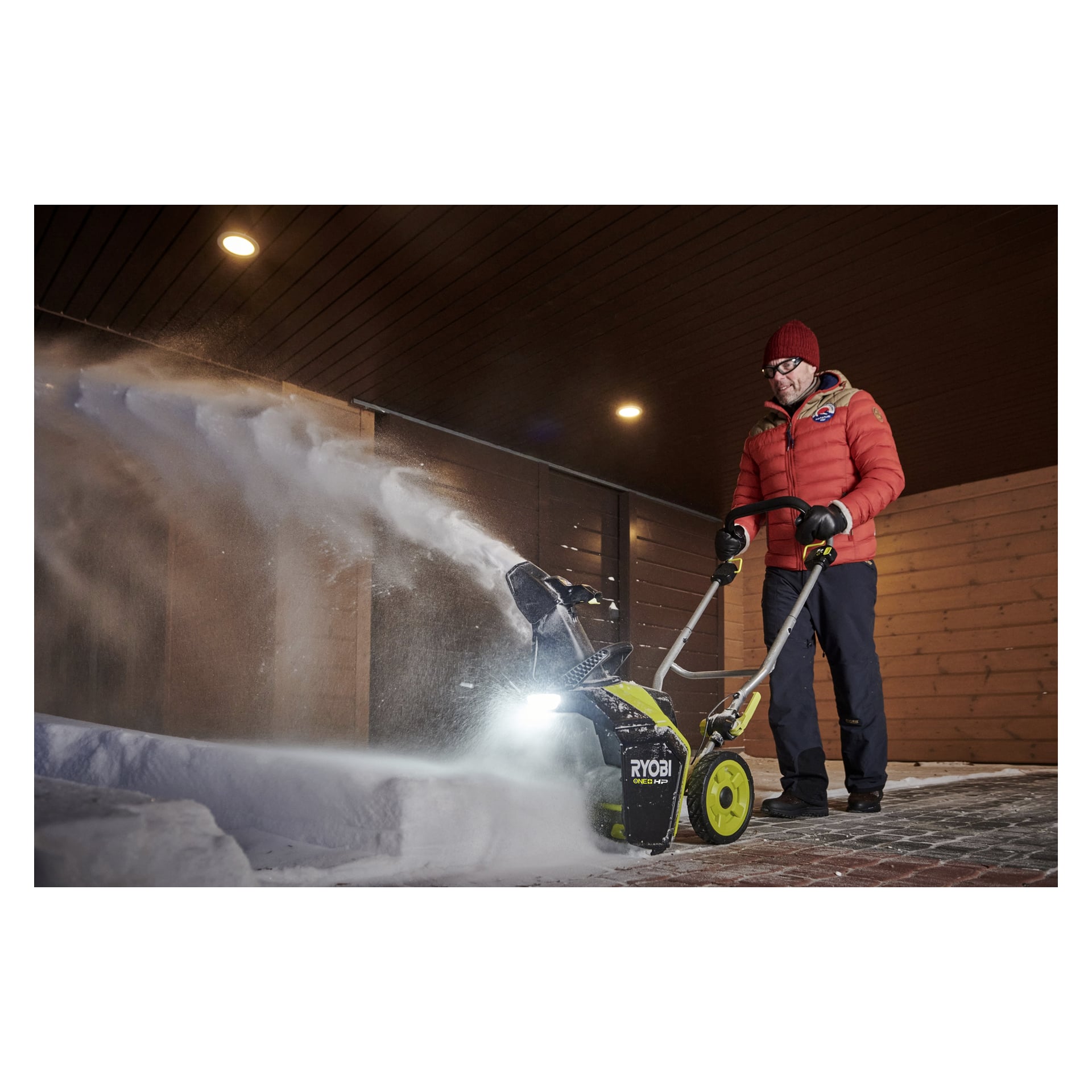 En person i röd jacka, mörka byxor och röd hatt kör en Ryobi-snöslunga på en snötäckt tegelstig under ett trätak.