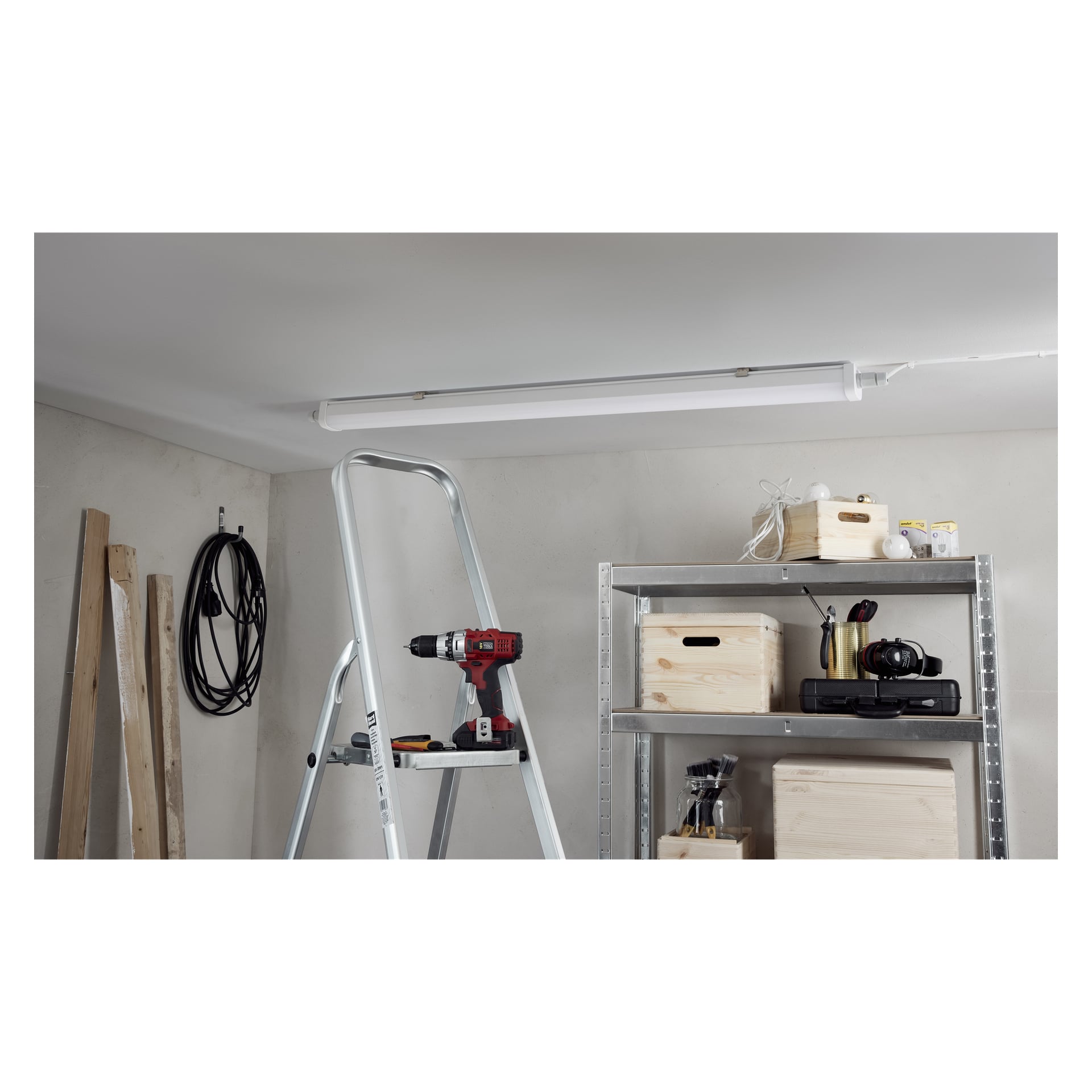 Eine Trittleiter ist unter einer Deckenlampe positioniert und hält eine rot-schwarze Bohrmaschine. In der Nähe befinden sich Kabel, Holzbretter und Regale mit Kisten.