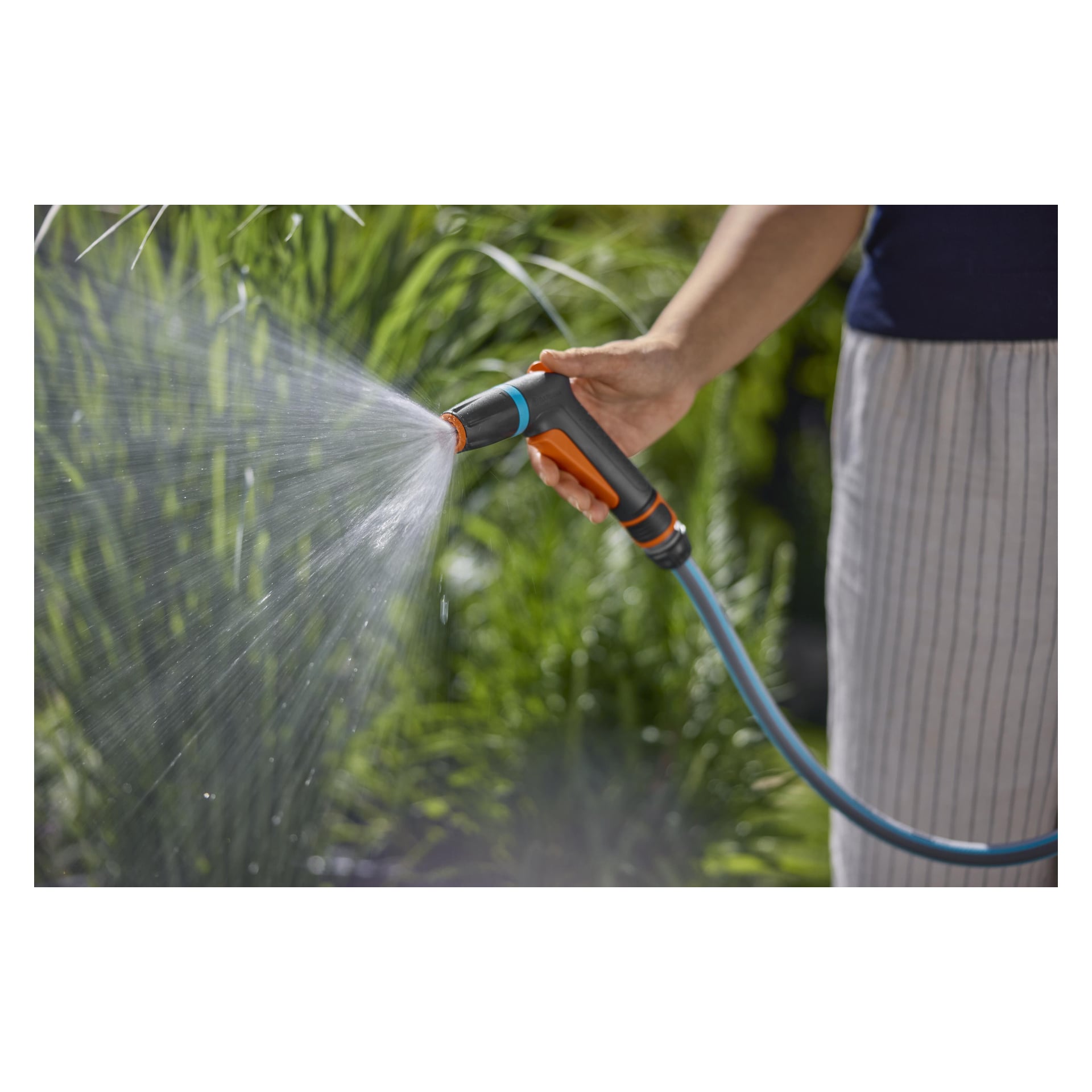 Jemand gießt Außenpflanzen mit einem Gartenschlauch mit einer Sprühdüse, umgeben von üppigem Grün und mit Streifen.