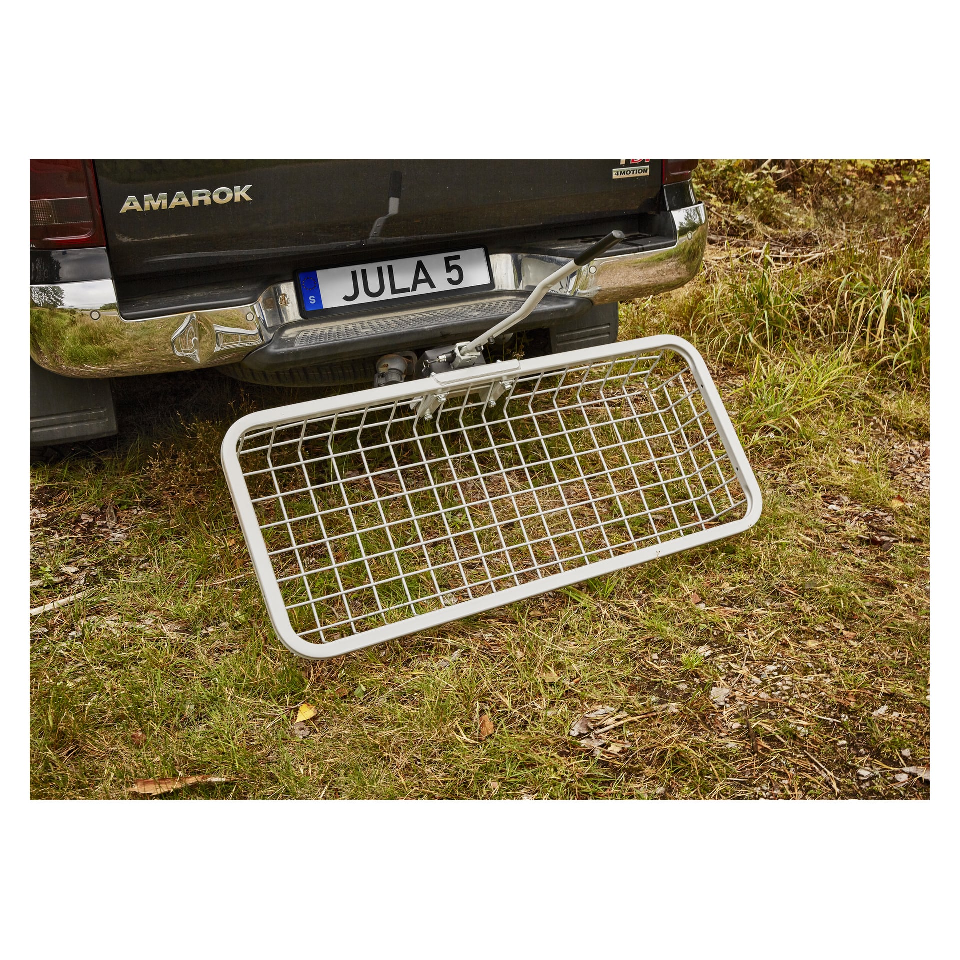 En metallgallerlastbärare är monterad på baksidan av en svart Amarok, delvis vilande på gräs och jord. Registreringsskylten syns.