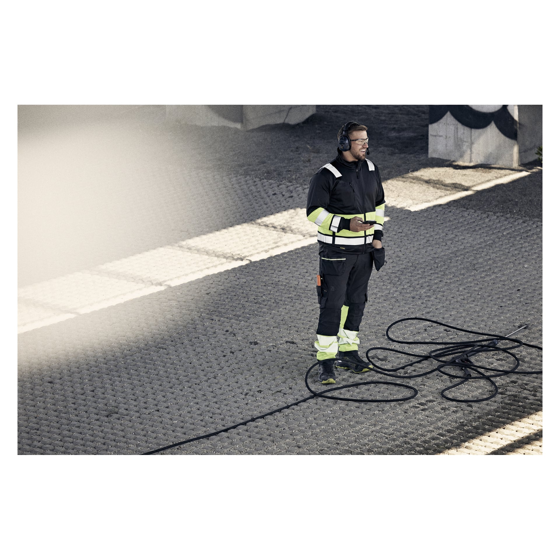 En man i en svart och neongul skyddsdräkt med hörlurar står på trottoaren med ringlade kablar och håller en apparat nära skuggor och betong.