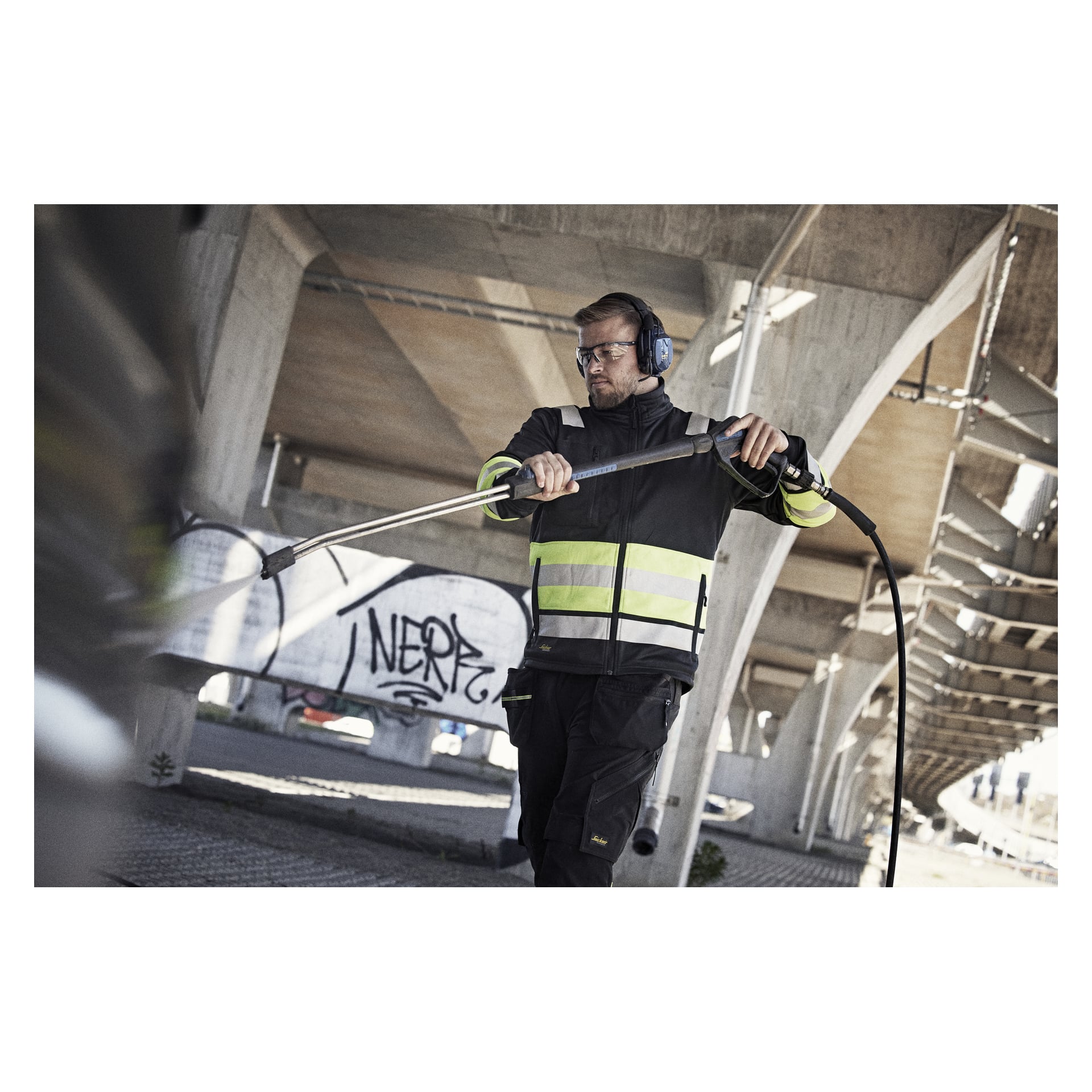 En man i skyddsutrustning och hörlurar använder en högtryckstvätt under en betongbro märkt med graffiti på stödpelarna.