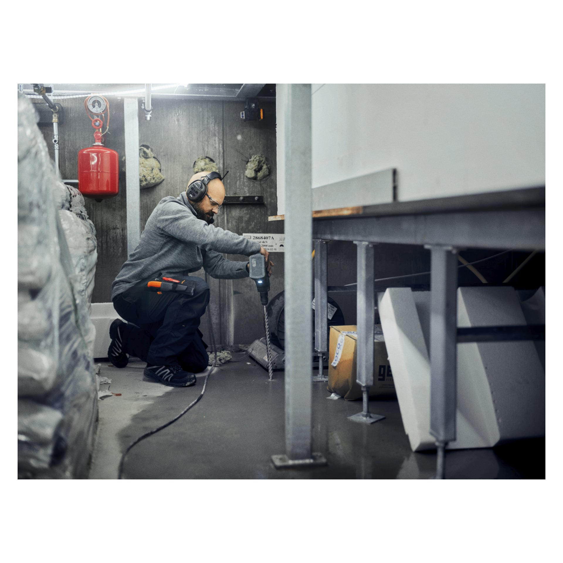 En person med hörlurar och skyddsglasögon knäböjer på betong och använder en borrmaskin bland isolering och verktyg på en byggarbetsplats.