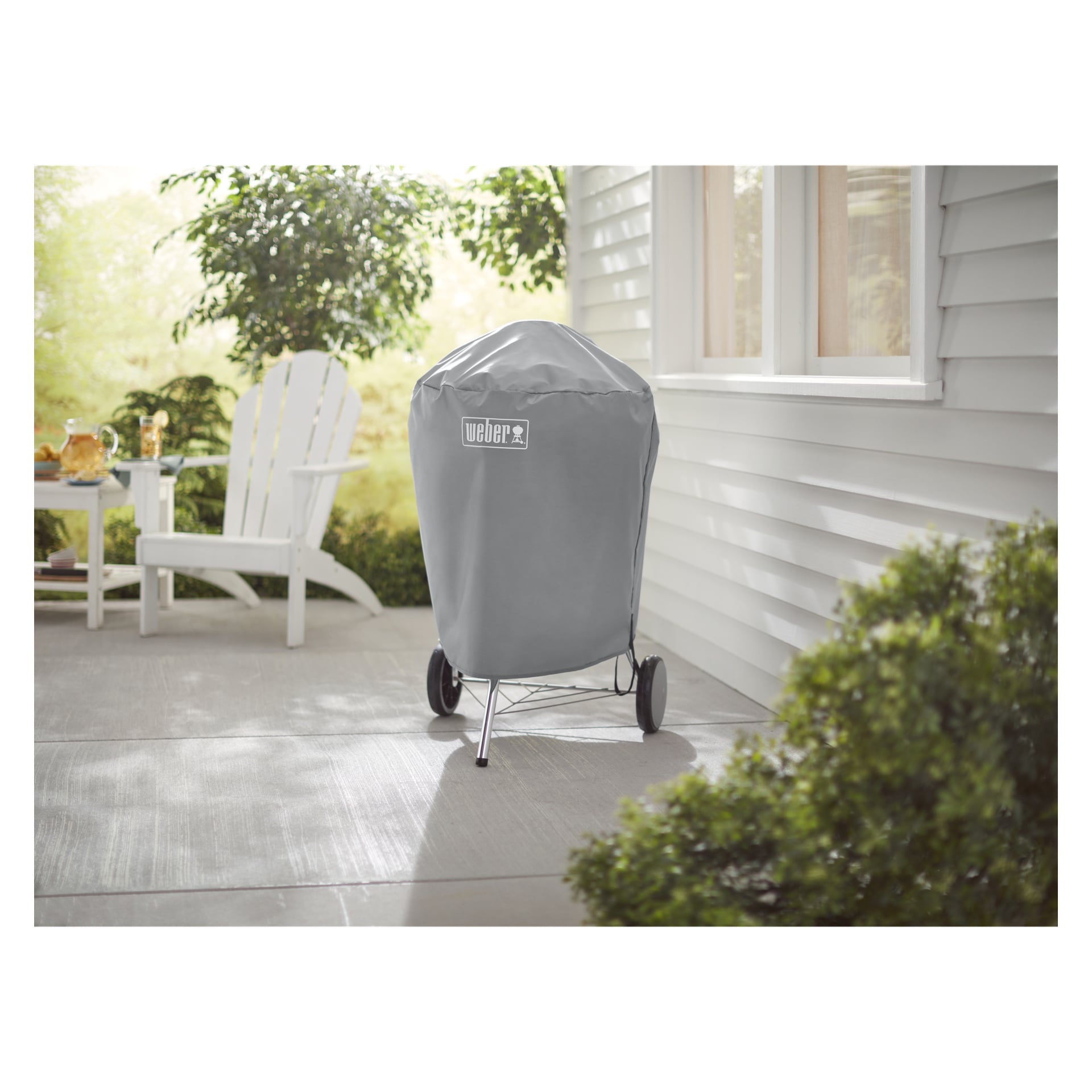 En overbygd Weber-grill står på en terrasse ved siden av et hvitt hus, med en Adirondack-stol og et lite bord omgitt av grøntområder.