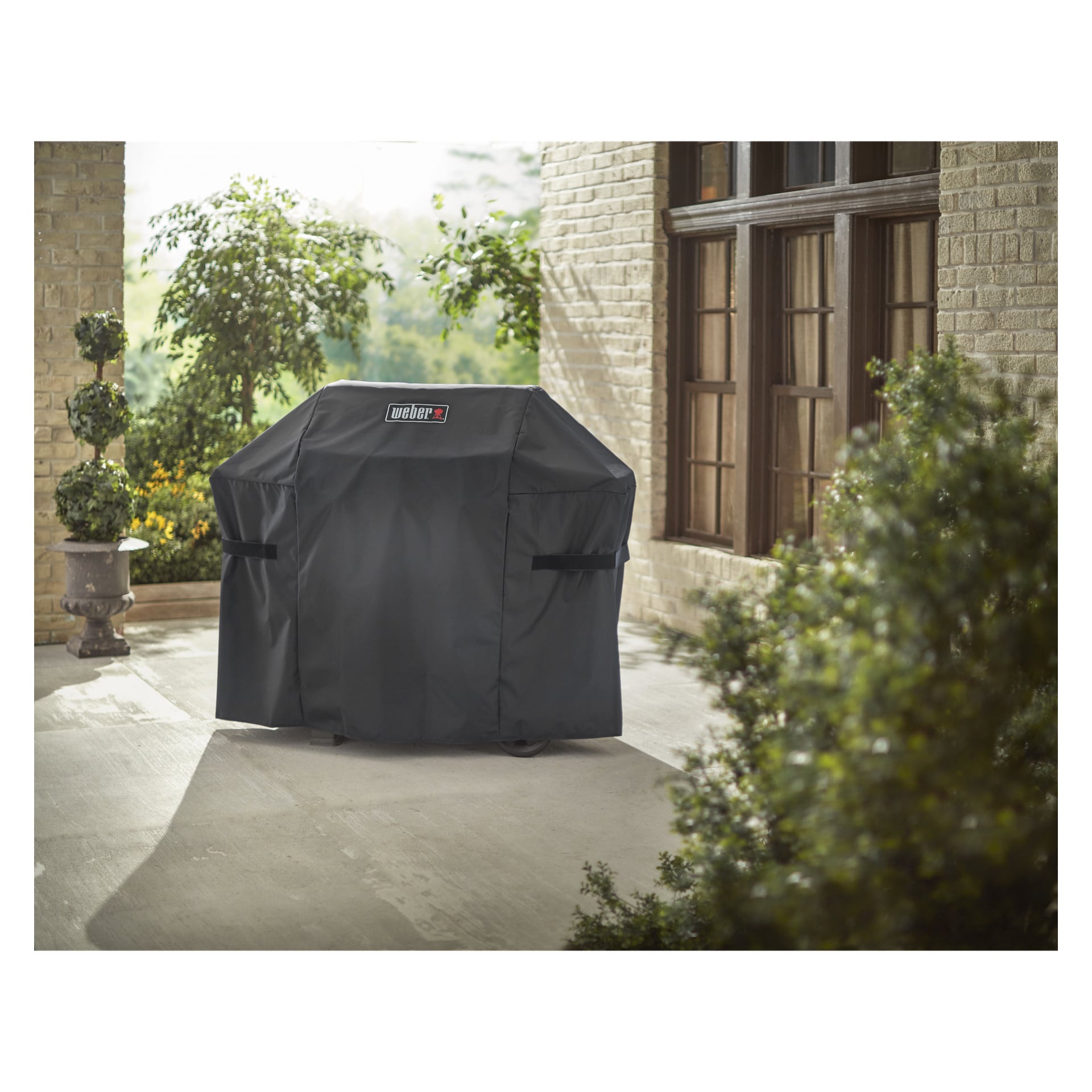 Eine schwarze Weber-Grillabdeckung steht auf einem Grill auf einer Terrasse mit grünen Topfpflanzen, neben einem hellen Backsteinhaus mit großen Fenstern.