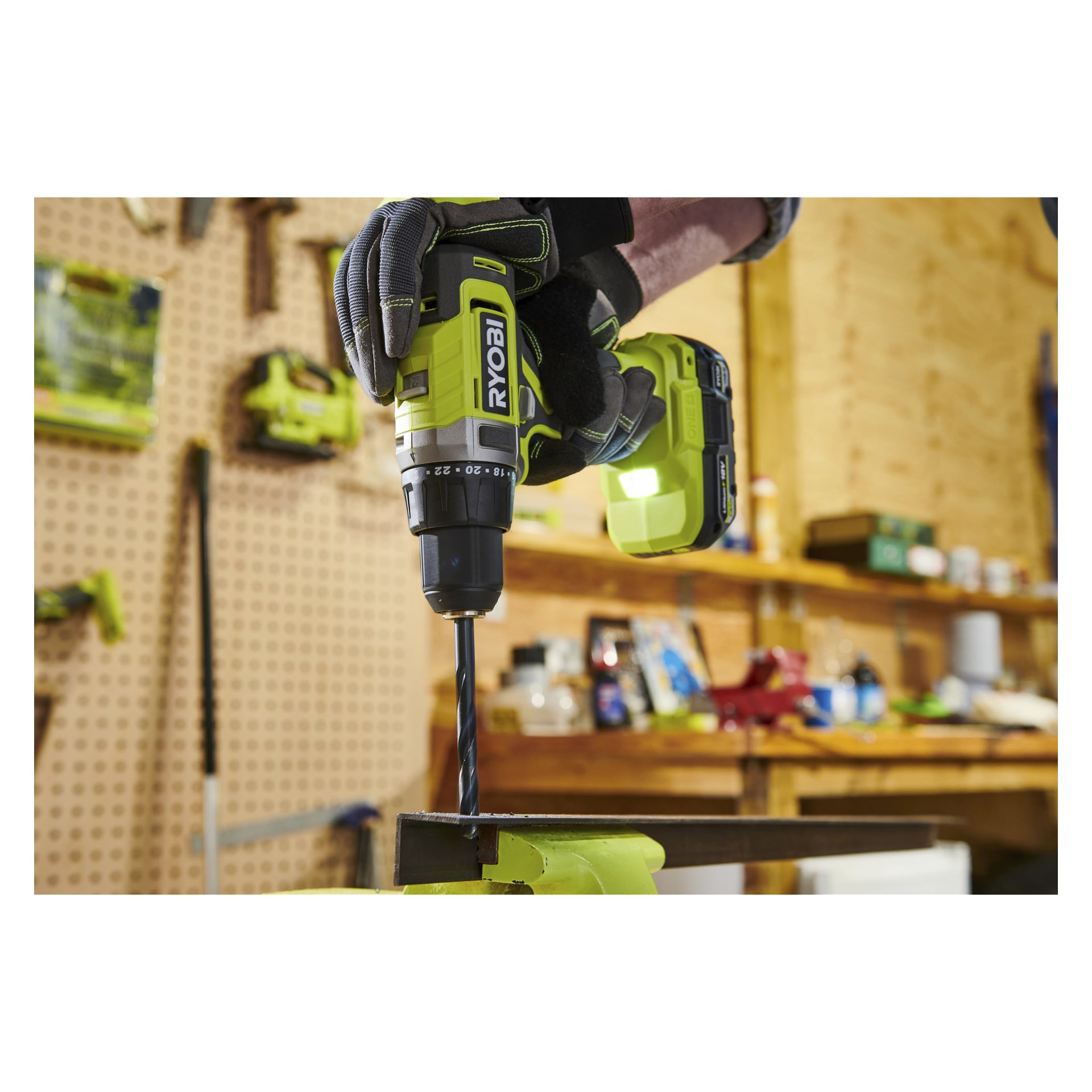 En hanskekledd hånd bruker en Ryobi trådløs drill til å bore i metall, omgitt av verkstedhyller og diverse verktøy i bakgrunnen.