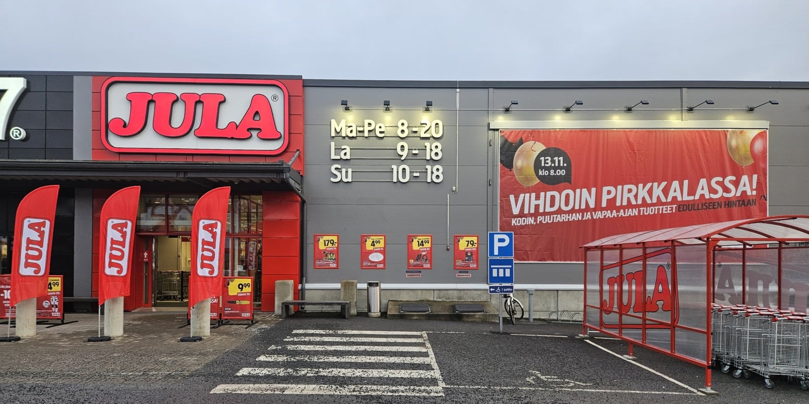 Jula-myymälän ulkotiloissa punaiset kyltit, neljä punaista lippua, myymälän aukioloajat seinällä, mainosbanneri, kärryt suojassa, märkä parkkipaikka.