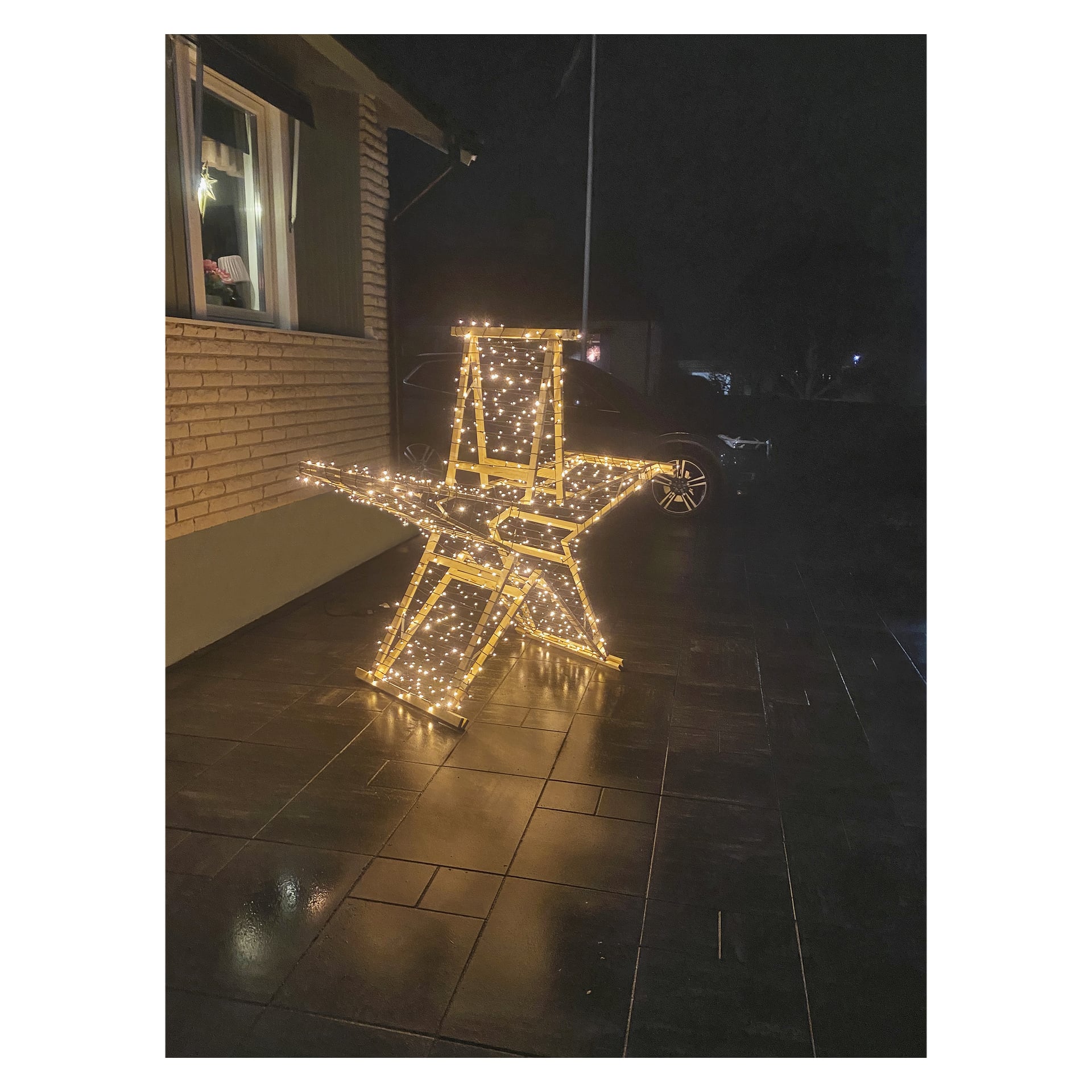 En trästege med stjärnformade ljusslingor står på våta kakel nära ett tegelhus, upplyst på natten.