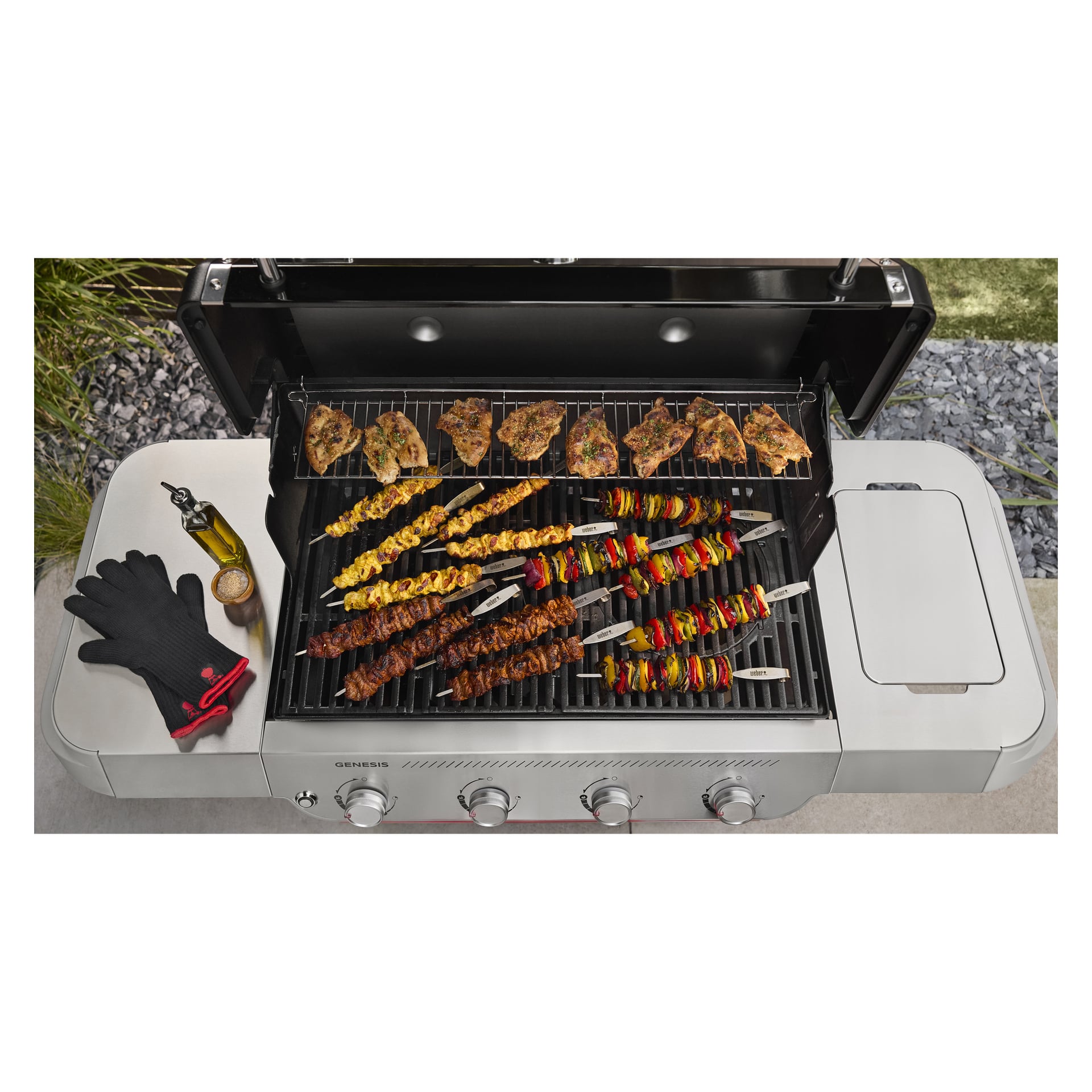 Ujęcie z góry na grill gazowy, w którym na bocznej półce znajdują się szaszłyki mięsa i warzyw, grillowany kurczak, czarne rękawice grillowe, miska do przypraw oraz butelka oleju.