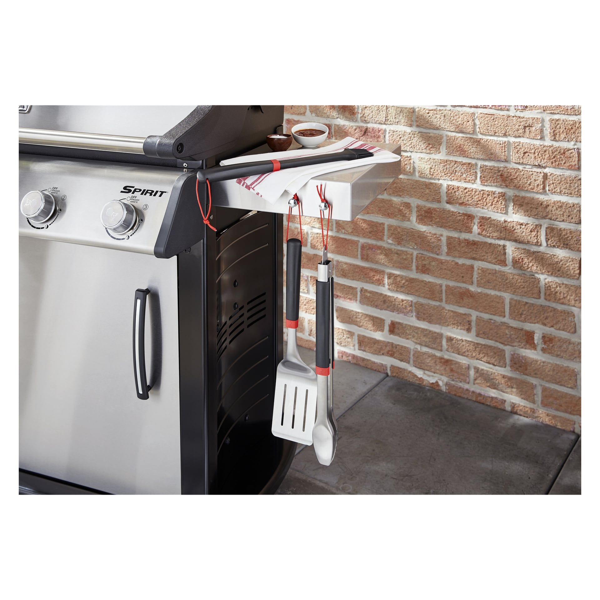 En rostfri gasgrill med två rattar står vid en tegelvägg. Sidohyllan har en vit bricka, randig handduk, såsskål och bestick.